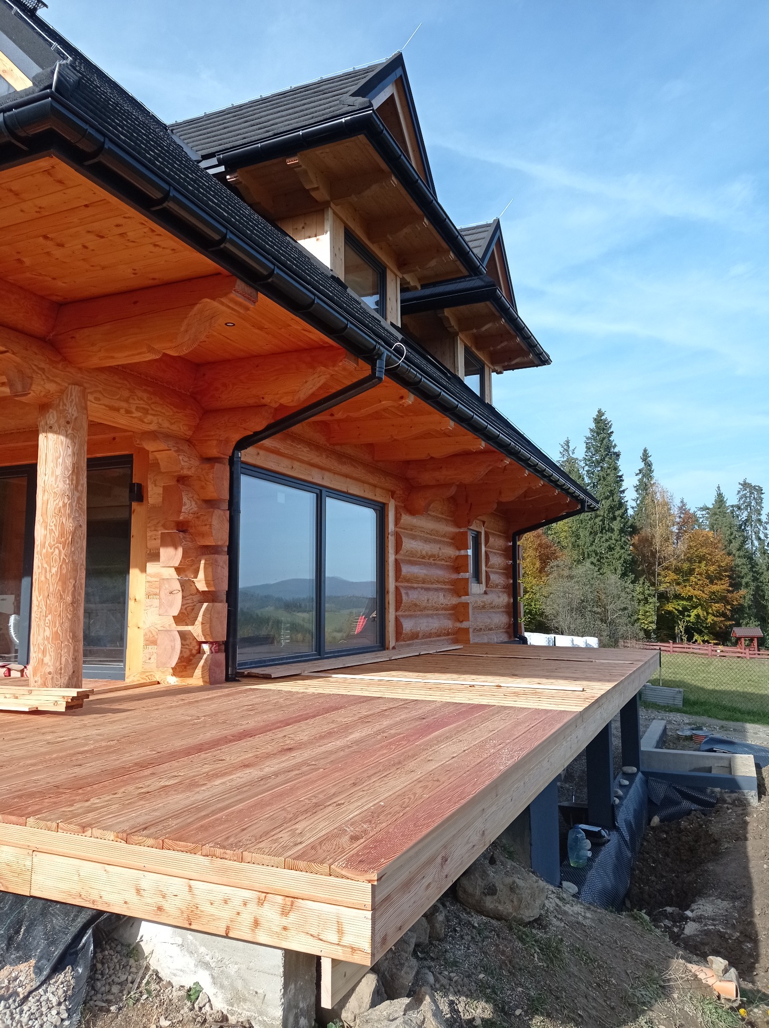 Drewniany dom z bali z dużym tarasem w trakcie budowy, widok na góry w tle, czarne rynny i dachówka, elementy konstrukcyjne tarasu jeszcze nie wykończone.