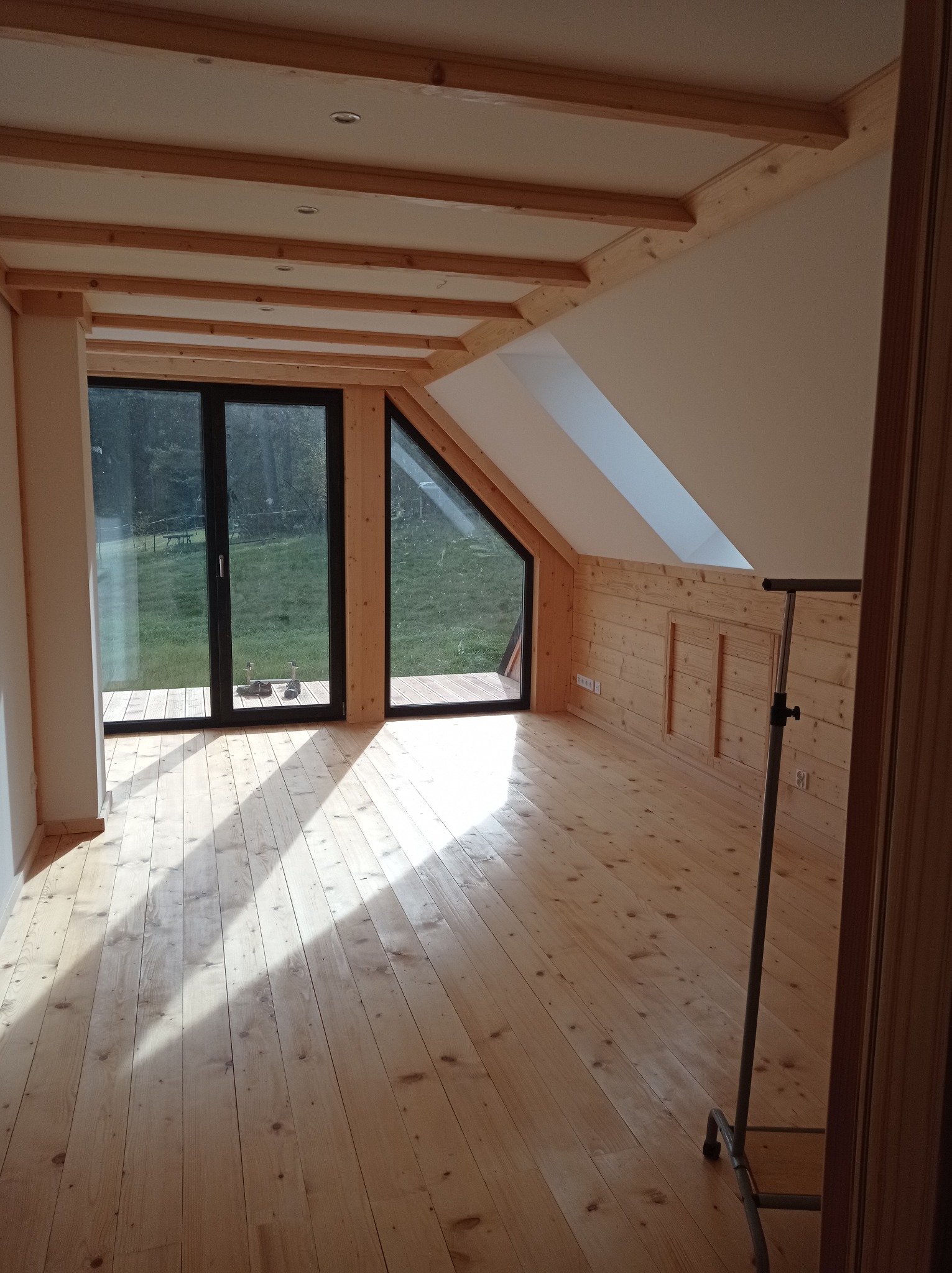 Jasne, przestronne poddasze z drewnianą podłogą i belkami stropowymi, duże okna z widokiem na zieleń, częściowo wykończone drewnianą boazerią, w rogu widoczny wieszak na ubrania.