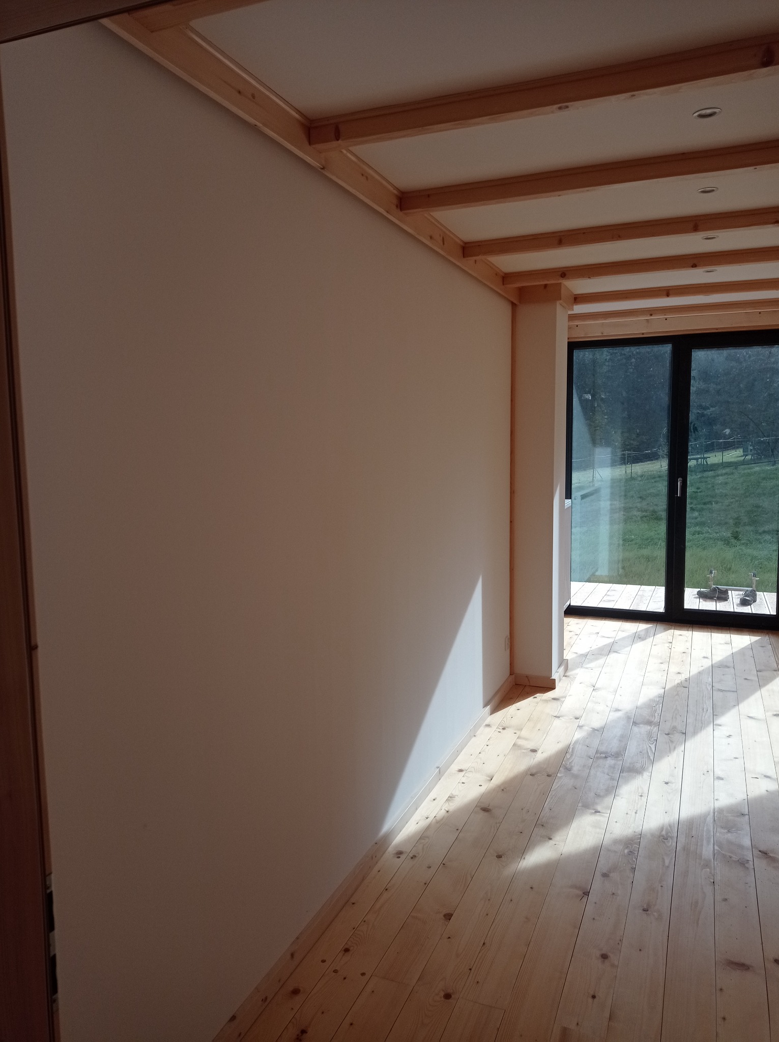 Jasne wnętrze z drewnianą podłogą i sufitem z belek, świeżo pomalowane ściany, duże okno z widokiem na zieleń na zewnątrz.