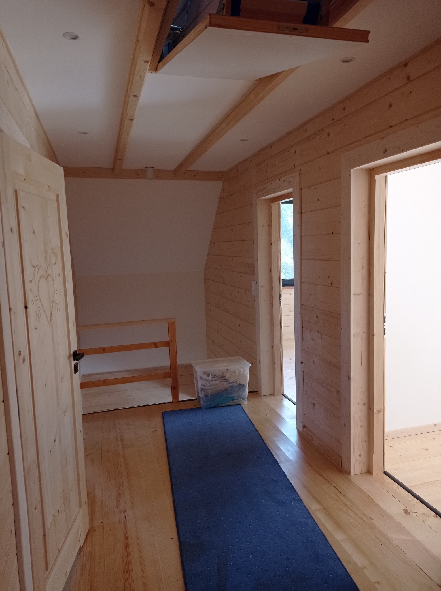 Drewniane wnętrze poddasza z widoczną otwartą klapą na strych, jasnymi ścianami z boazerii, drewnianą podłogą i niebieskim dywanikiem.
