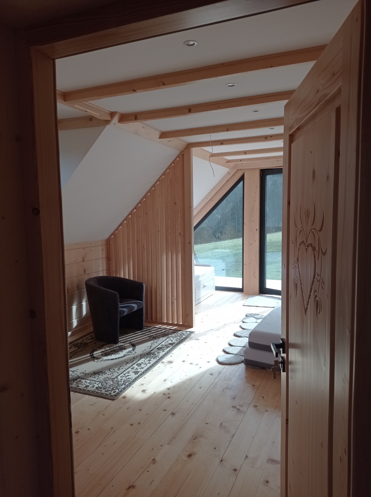 Drewniane wykończenie poddasza z widoczną konstrukcją stropu, krzesłem i dywanem, widok zza otwartych drzwi z dekoracyjnym wzorem.
