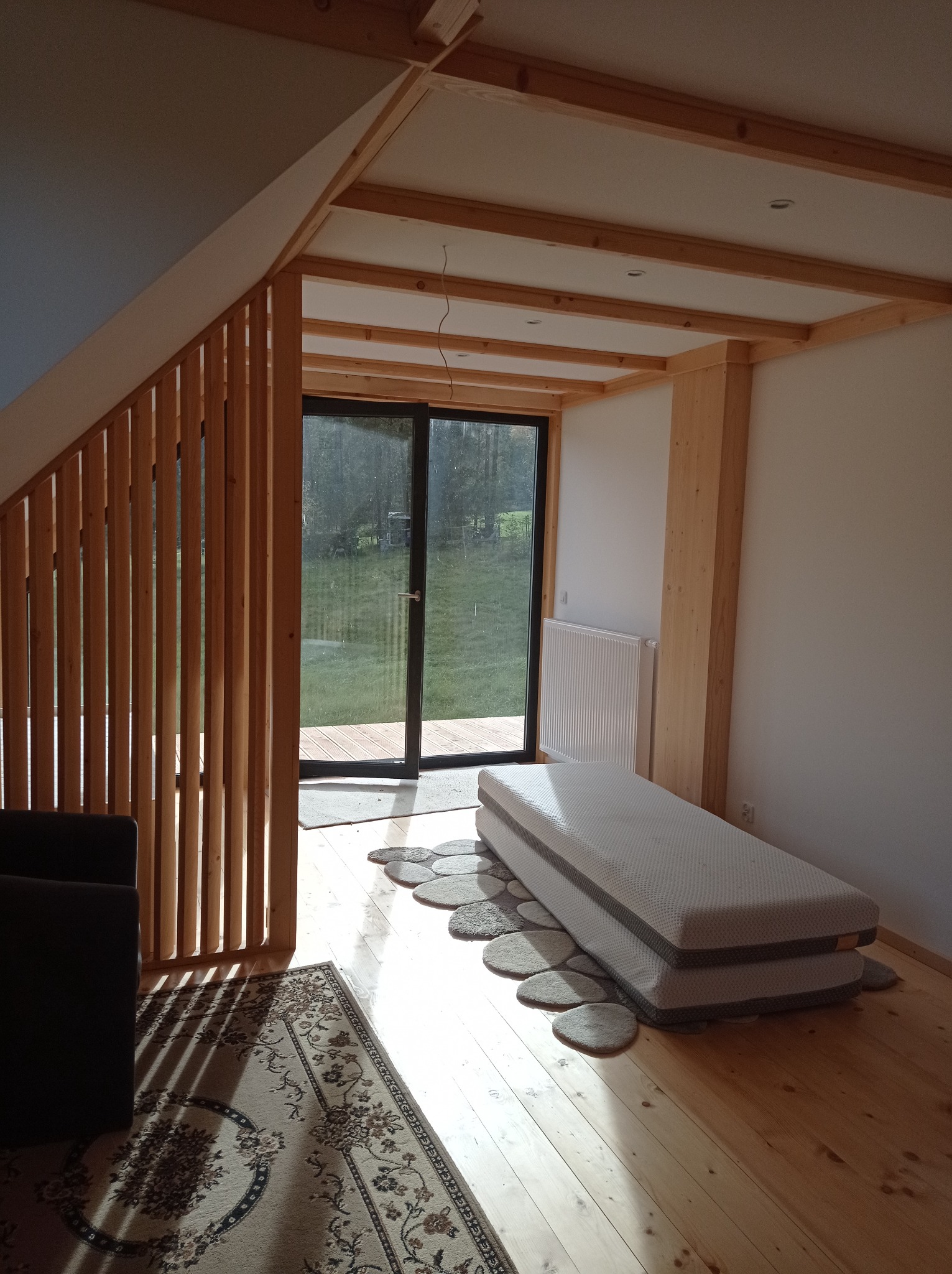 Surowe wnętrze poddasza z drewnianą konstrukcją stropu, ażurową ścianką działową, dużym oknem tarasowym i materacem na podłodze, w trakcie wykańczania.