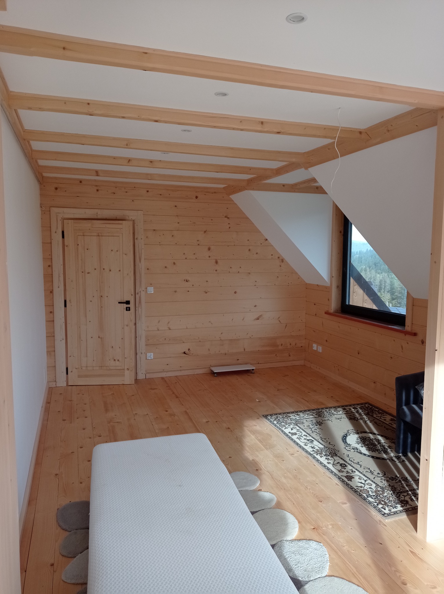 Pokój na poddaszu wykończony drewnem, z widocznymi belkami stropowymi, oknem dachowym, drewnianą podłogą i drzwiami, częściowo umeblowany materacem i dywanem.