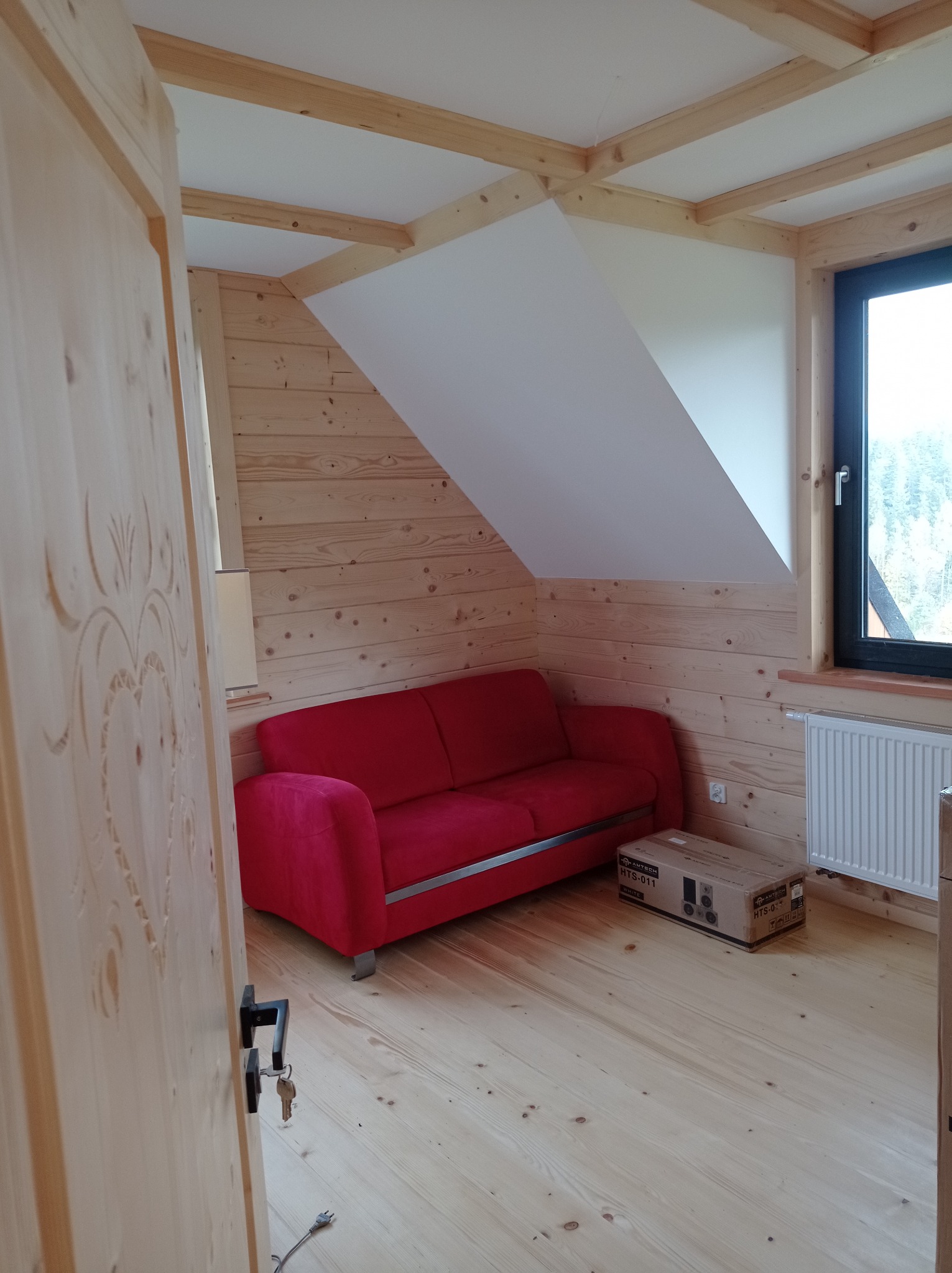 Pokój na poddaszu wykończony boazerią z jasnego drewna, z czerwonym narożnikiem, białym grzejnikiem i oknem, widok zza otwartych, zdobionych drzwi.