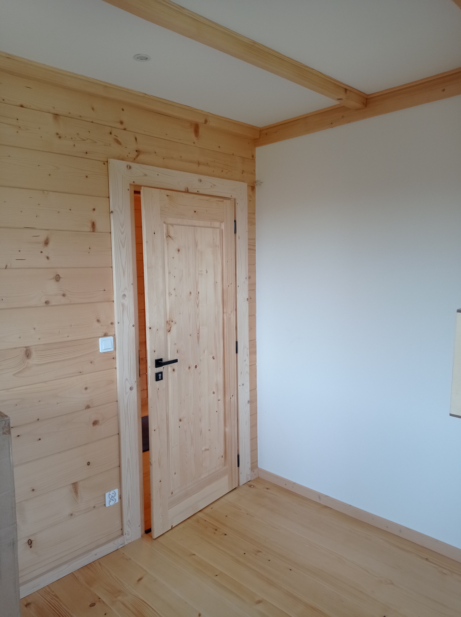 Wnętrze pokoju wykończonego drewnem: boazeria, podłoga i drzwi z jasnego drewna, kontrastujące z białą ścianą i czarną klamką, widoczne gniazdko elektryczne i włącznik światła.