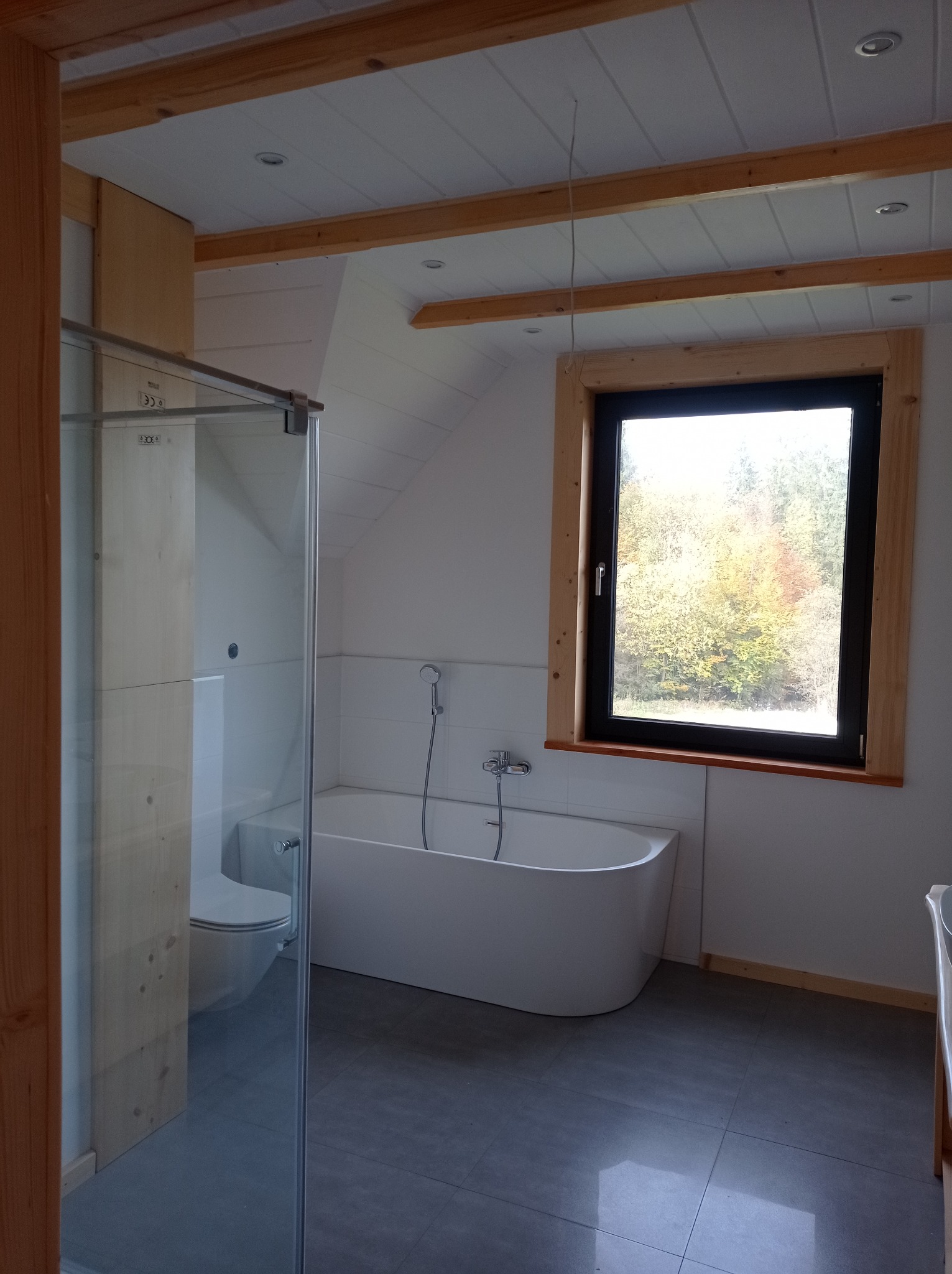 Nowoczesna łazienka z wolnostojącą wanną, kabiną prysznicową ze szklanymi drzwiami, toaletą w zabudowie, szarą podłogą i oknem z widokiem na drzewa.