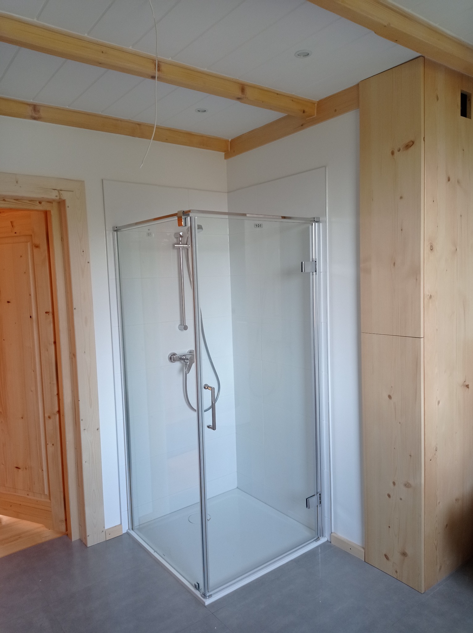 Nowoczesna kabina prysznicowa z chromowanymi okuciami w jasnej łazience z drewnianymi elementami wykończeniowymi sufitu i drzwi, widoczna również szafa z surowego drewna.