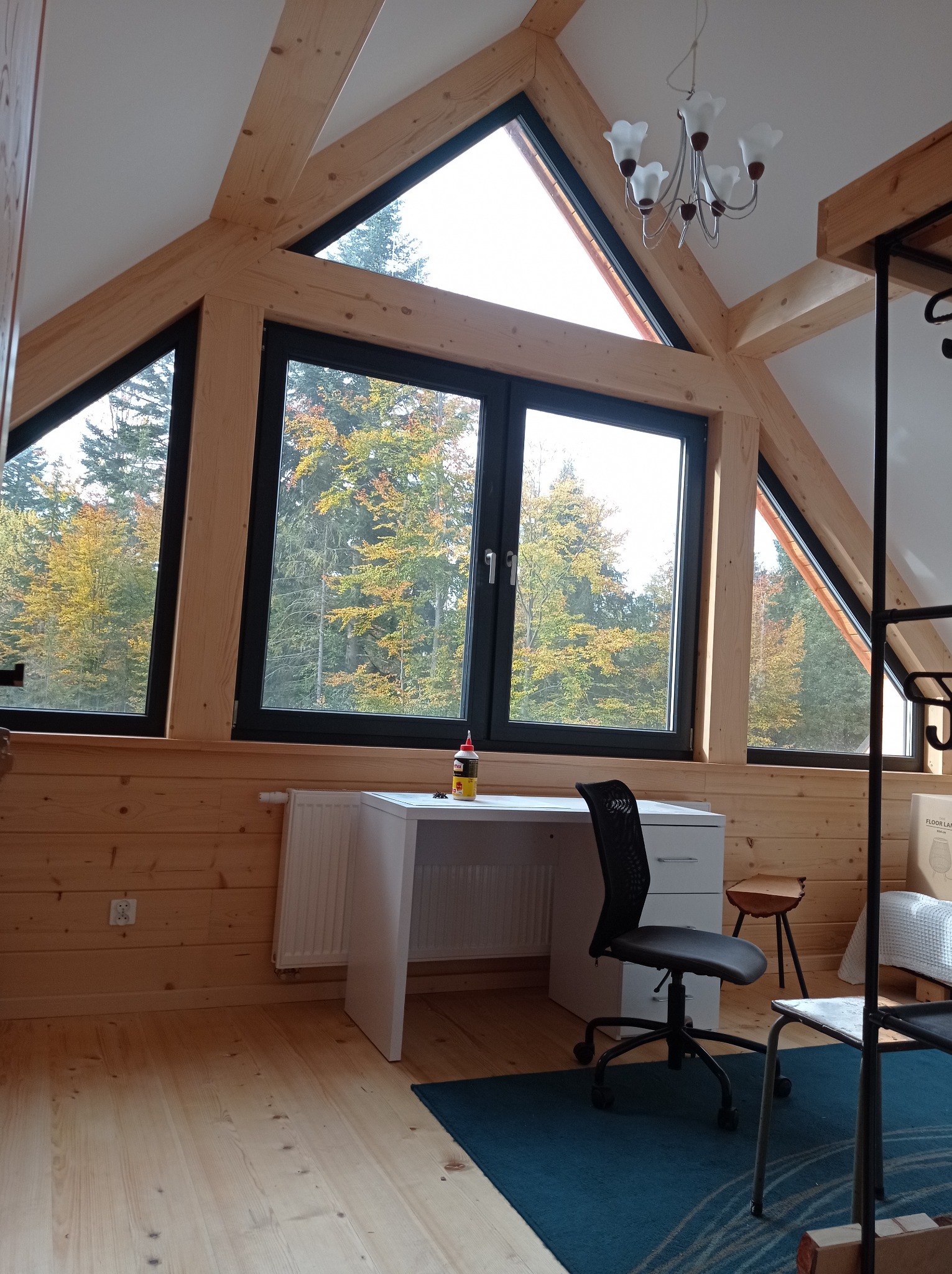 Pokój na poddaszu z drewnianą boazerią, oknami z widokiem na jesienny las, białym biurkiem i krzesłem biurowym na niebieskim dywanie.