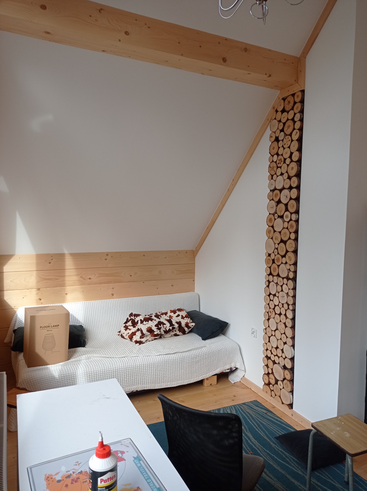 Pokój na poddaszu z drewnianą boazerią i dekoracyjnym panelem z przekrojów pni drzew, biała sofa z poduszkami, karton po lampie na podłodze, stół z klejem Pattex i mapa świata.