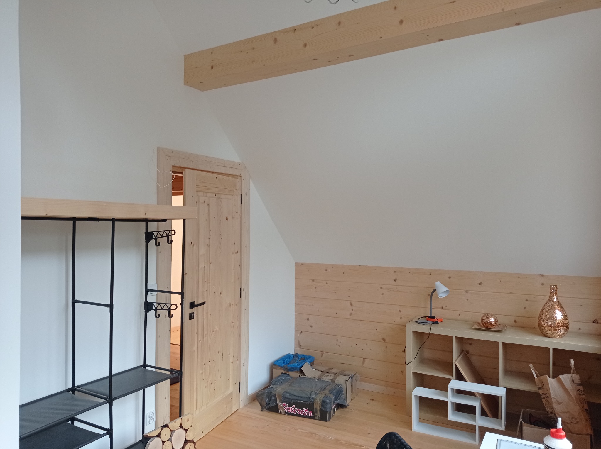 Pokój na poddaszu z drewnianą boazerią do połowy ściany, drewnianą belką stropową, drewnianą podłogą i drzwiami z surowego drewna. Widoczny regał z oświetleniem i dekoracjami, a także czarny regał...