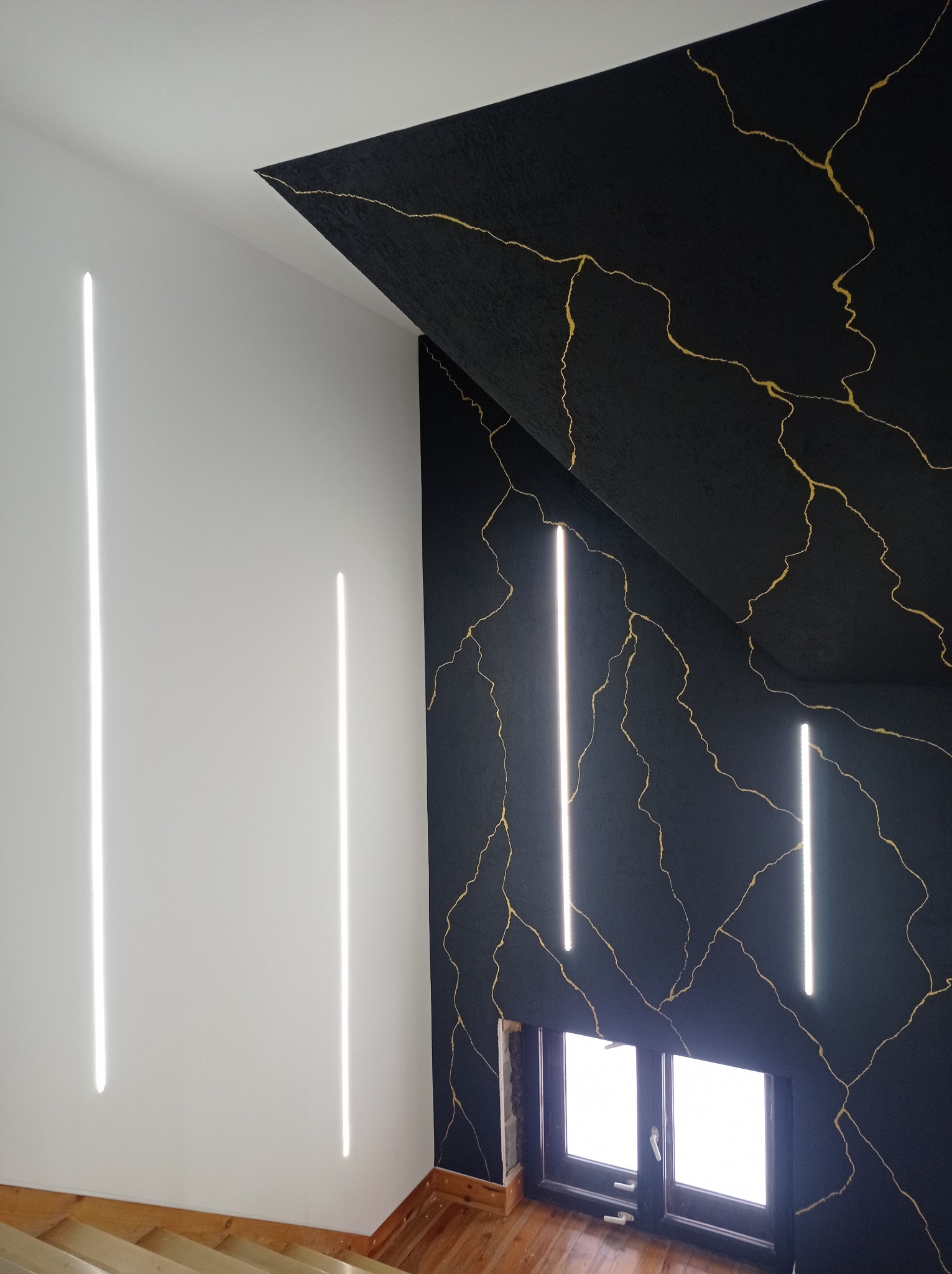 Nowoczesne wnętrze: czarna ściana z motywem złotych pęknięć, oświetlona pionowymi lampami LED, kontrastująca z białą ścianą i drewnianymi schodami.