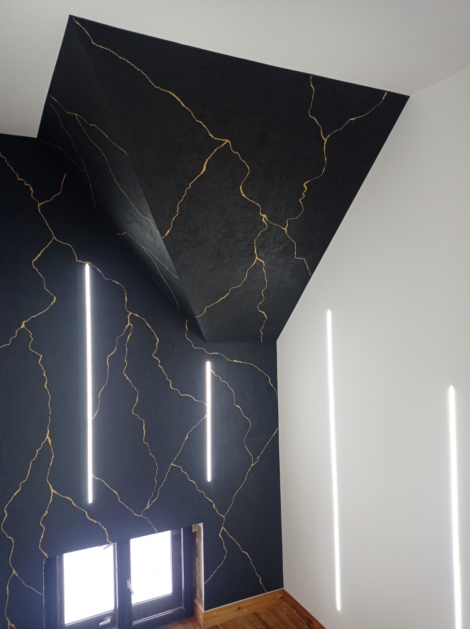 Czarno-złota ściana z efektem marmuru i pionowymi lampami LED, widok na okno i fragment drewnianej podłogi.