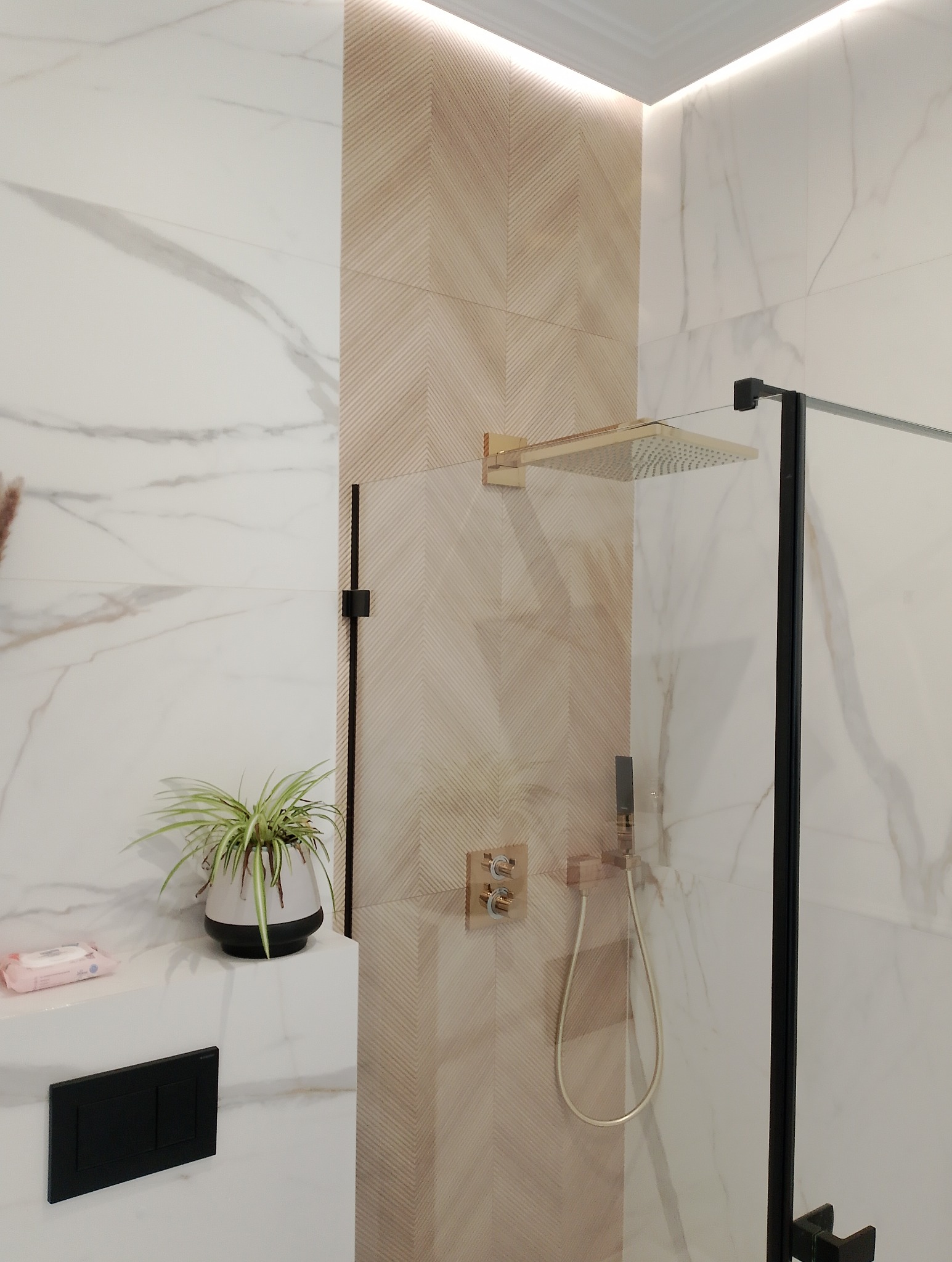 Nowoczesna łazienka z kabiną prysznicową typu walk-in, wykończona płytkami imitującymi marmur i drewno, z deszczownicą w kolorze złota i czarnymi elementami armatury, dopełniona rośliną doniczkową.
