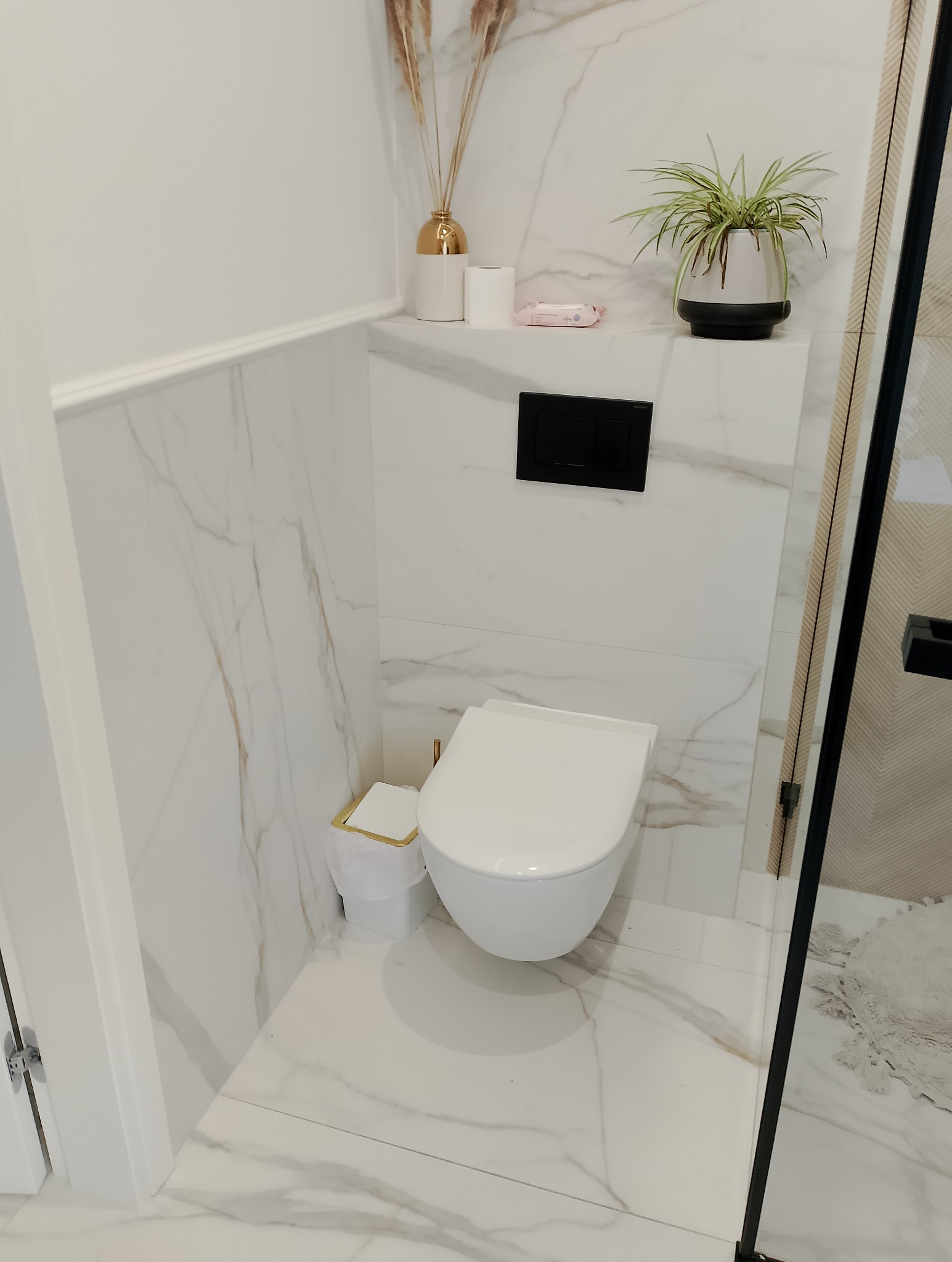Nowoczesna toaleta podwieszana z czarnym przyciskiem spłukującym, biała marmurowa okładzina ścienna i podłogowa, dekoracje w postaci suszonych traw w złotym wazonie oraz zielonej rośliny...