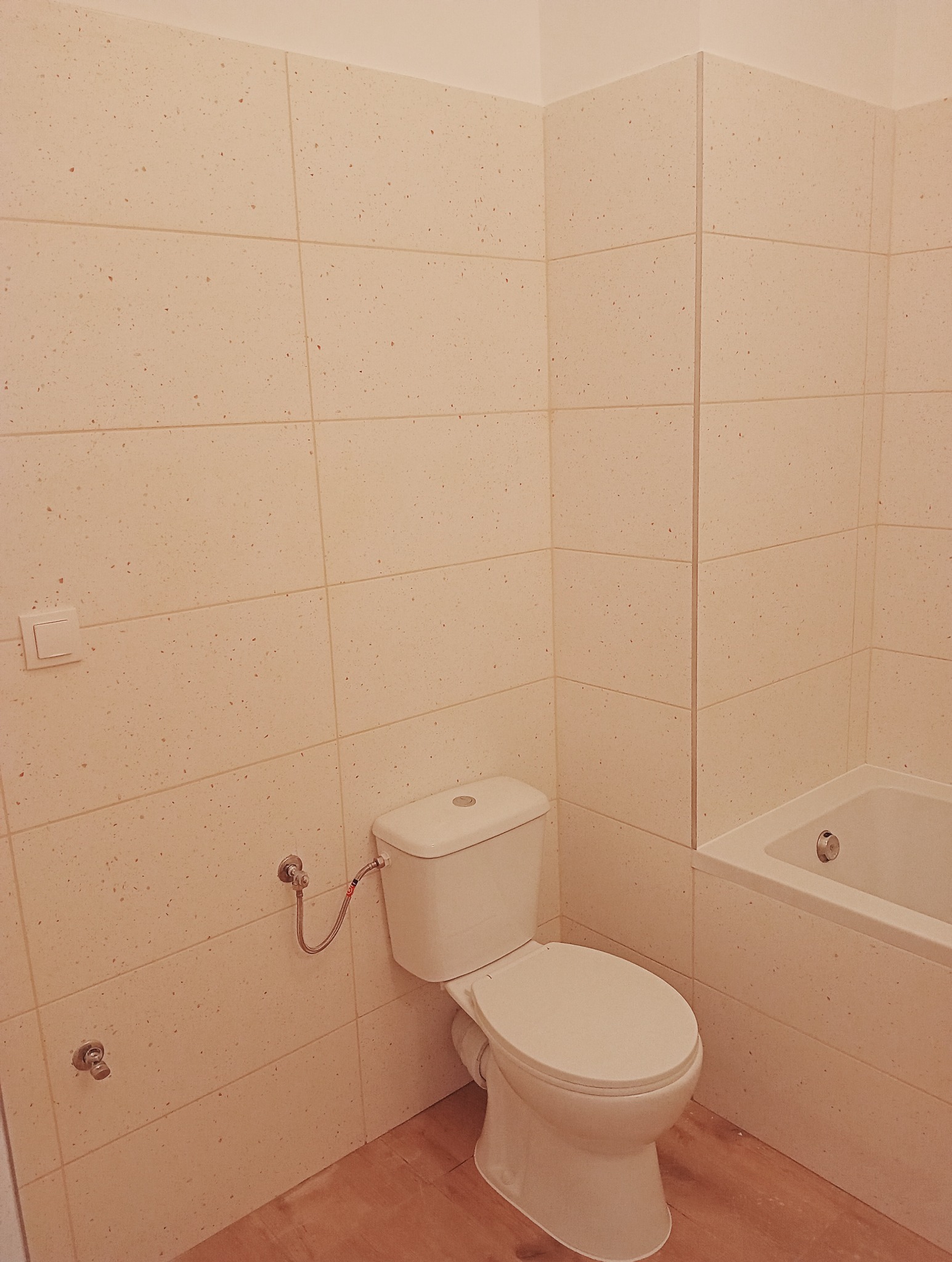 Jasna łazienka z beżowymi, prostokątnymi płytkami imitującymi kamień, widoczny biały sedes i fragment wanny.