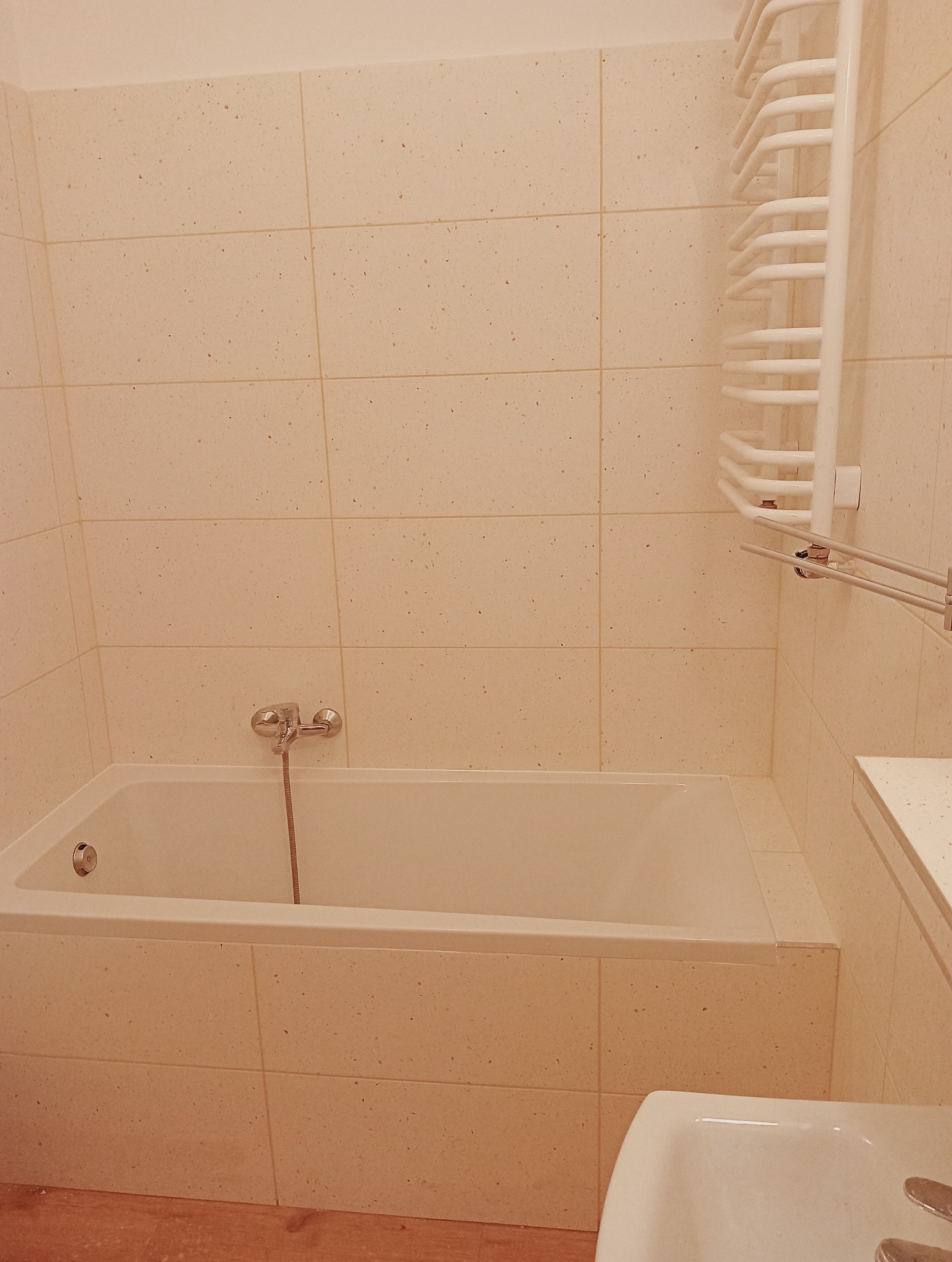 Jasna łazienka z wanną obłożoną płytkami w odcieniach beżu, na ścianie grzejnik drabinkowy i bateria wannowa z wężem.