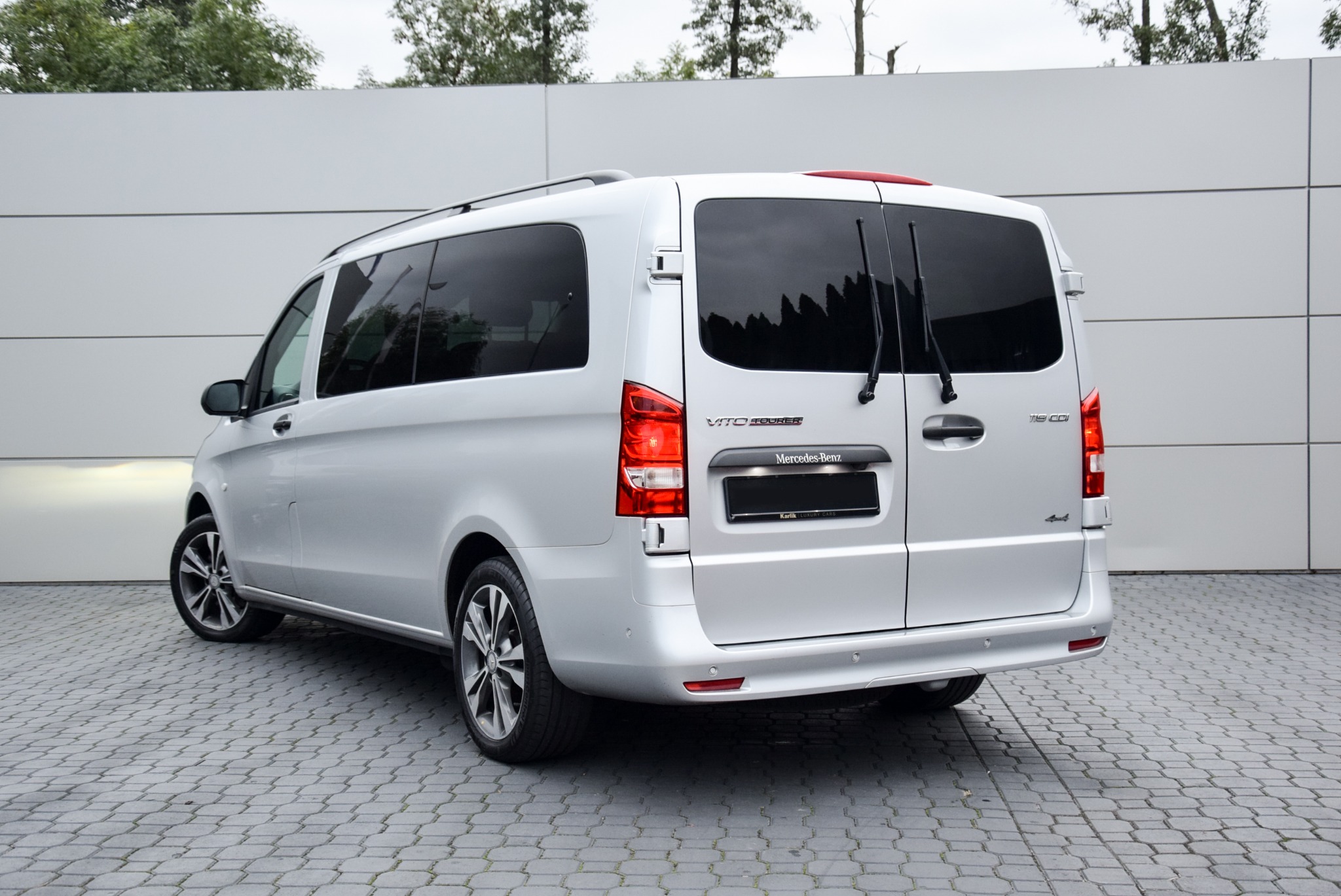 Srebrny Mercedes-Benz Vito Tourer z ciemnymi szybami, widok z tyłu na tle jasnoszarej ściany i kostki brukowej.