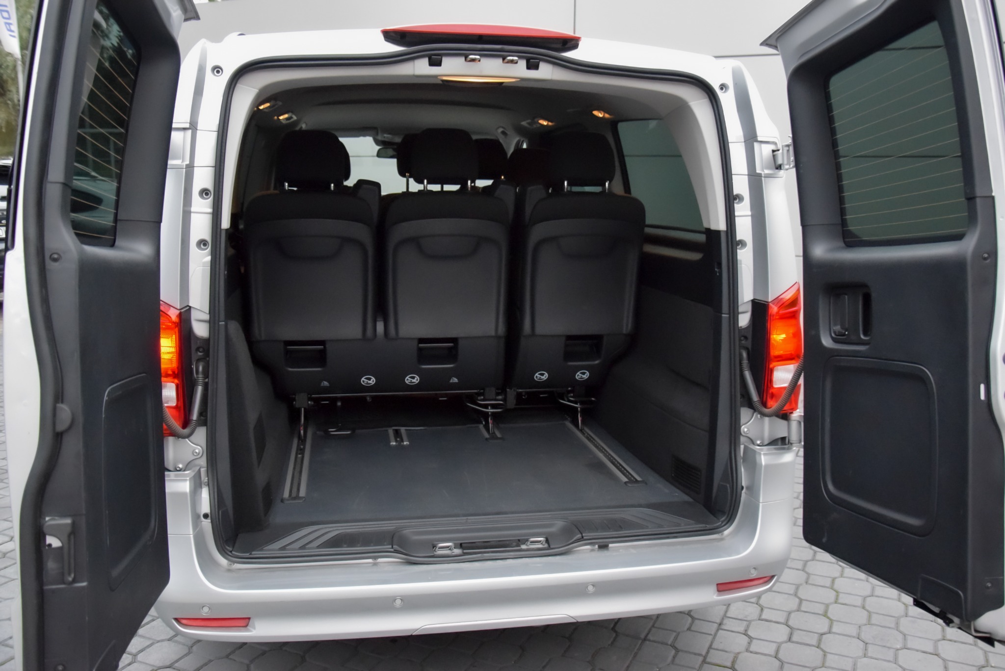 Wnętrze srebrnego busa z otwartymi drzwiami bagażnika, widoczne trzy rzędy czarnych foteli i przestrzeń bagażowa z szynami.