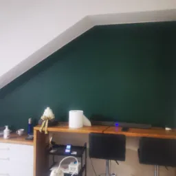 Poddasze po remoncie: zielona ściana w skośnym pomieszczeniu, białe meble z szufladami pod skosem, drewniany blat, dekoracje, dwa krzesła barowe i białe drzwi.