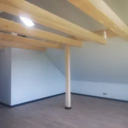 Poddasze z drewnianą konstrukcją więźby dachowej, świeżo wykończone ściany pomalowane na biało, widoczne gniazdka elektryczne i lampa wisząca na kablu.