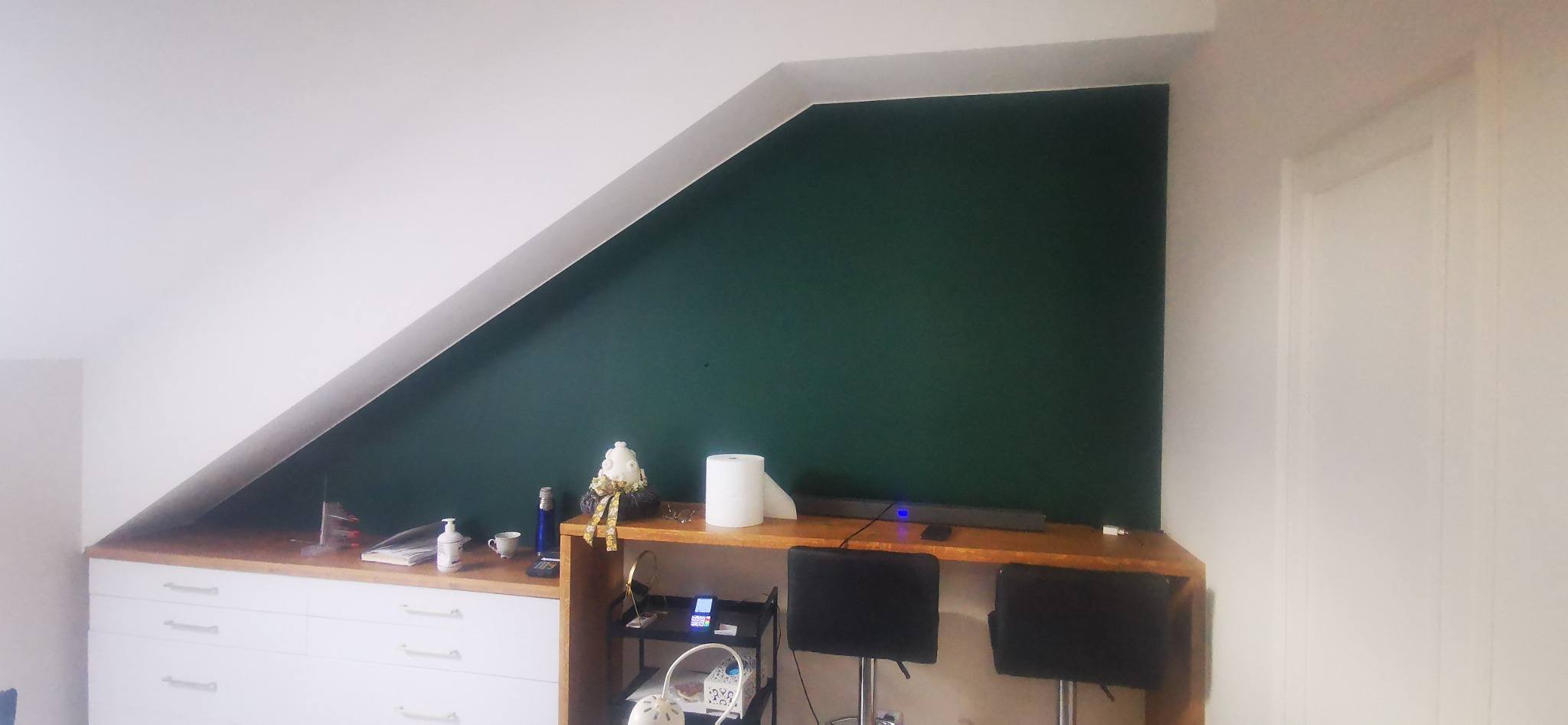 Poddasze po remoncie: zielona ściana w skośnym pomieszczeniu, białe meble z szufladami pod skosem, drewniany blat, dekoracje, dwa krzesła barowe i białe drzwi.