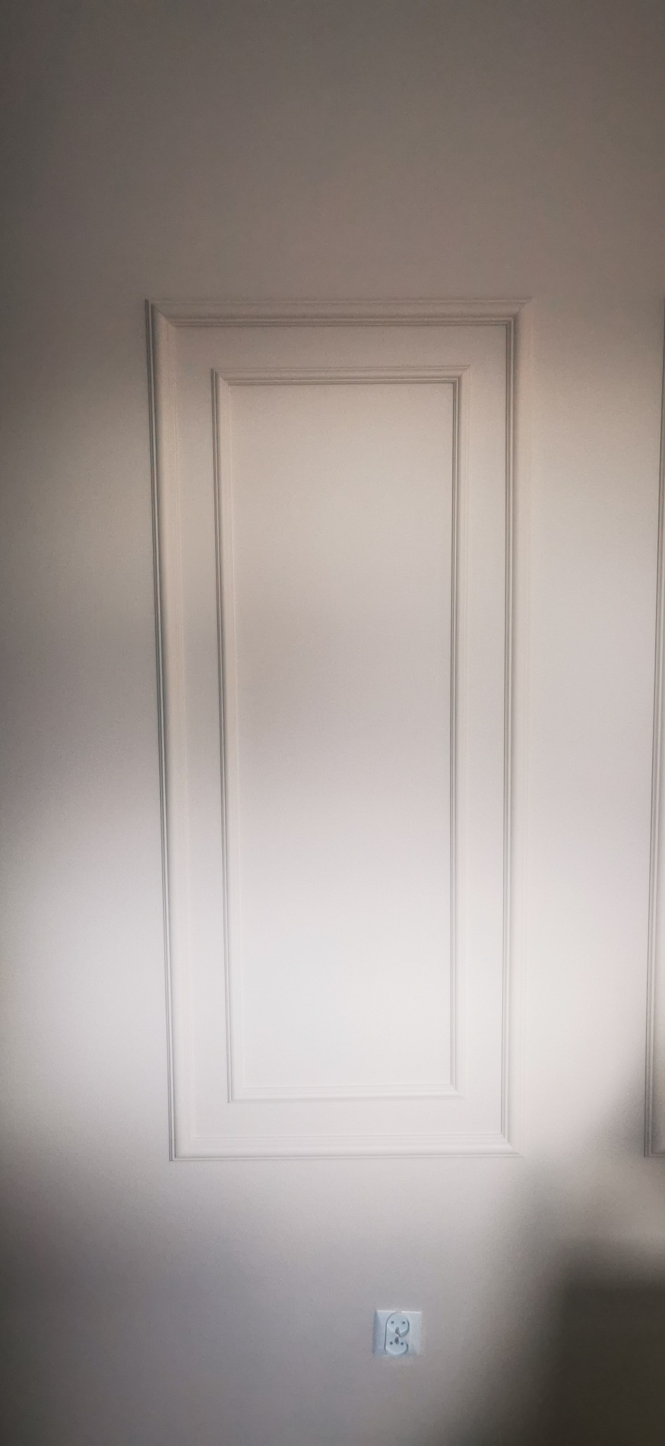 Biała ściana z dekoracyjnymi listwami i gniazdkiem elektrycznym w dolnej części.