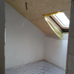 Podłogowe ogrzewanie w pokoju na poddaszu z oknem dachowym zabezpieczonym folią, ściany przygotowane do malowania, sufit z płyty OSB z wystającym kablem.