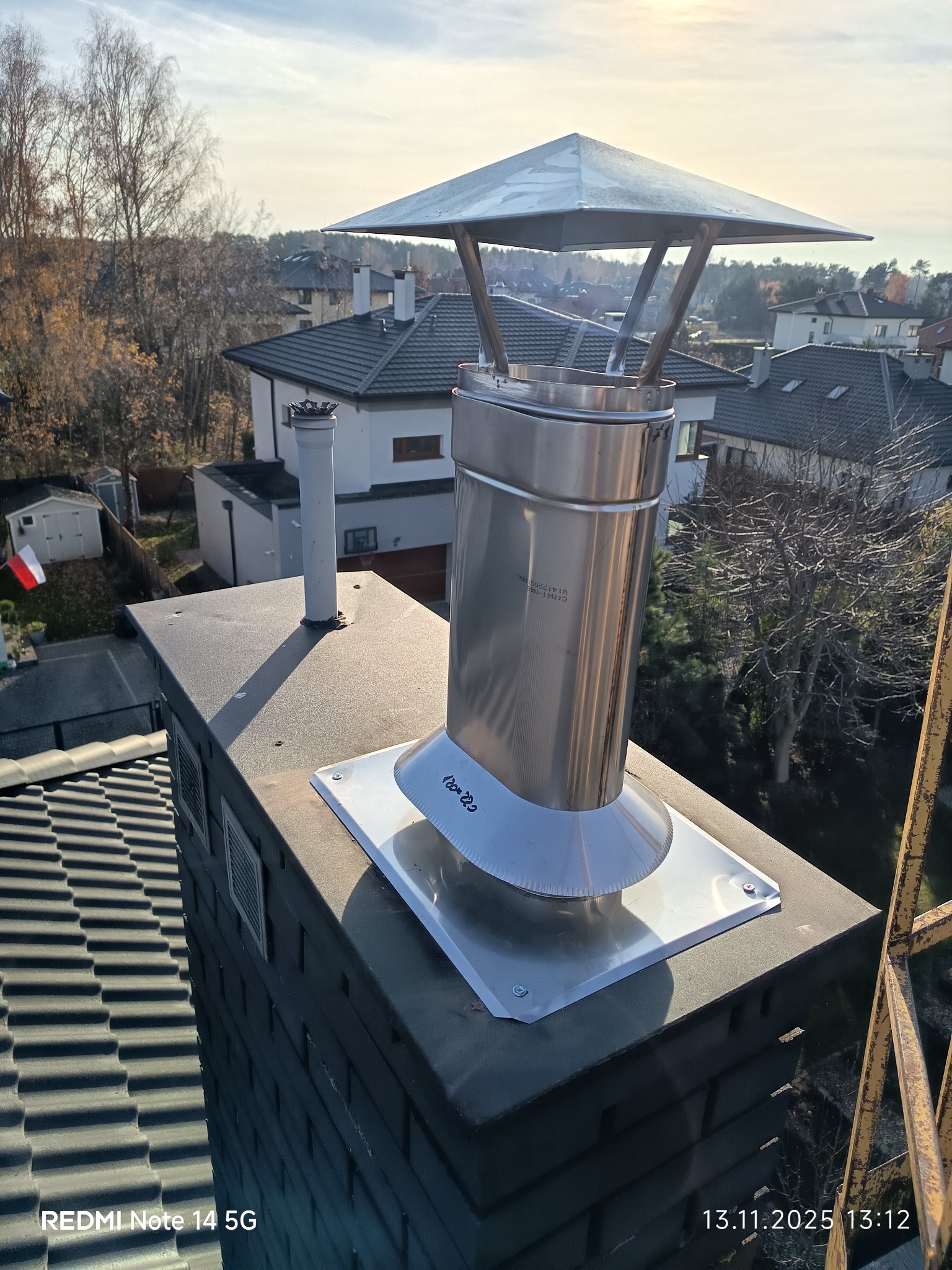 Montaż wkładu kominowego ze stali nierdzewnej na dachu domu jednorodzinnego, widok z góry, w tle zabudowa mieszkalna i drzewa. Słoneczny dzień.