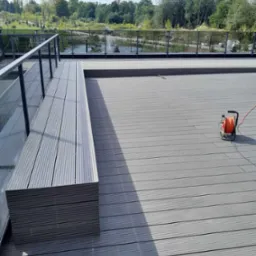 Szary taras kompozytowy z widokiem na park, z zamontowaną ławką i szklaną balustradą, pomarańczowy przedłużacz leży na podłodze.