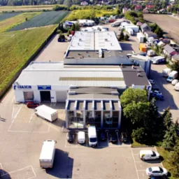 Widok z lotu ptaka na budynek firmy z zaparkowanymi busami i ciężarówkami, w tle pola uprawne i zabudowa mieszkalna.