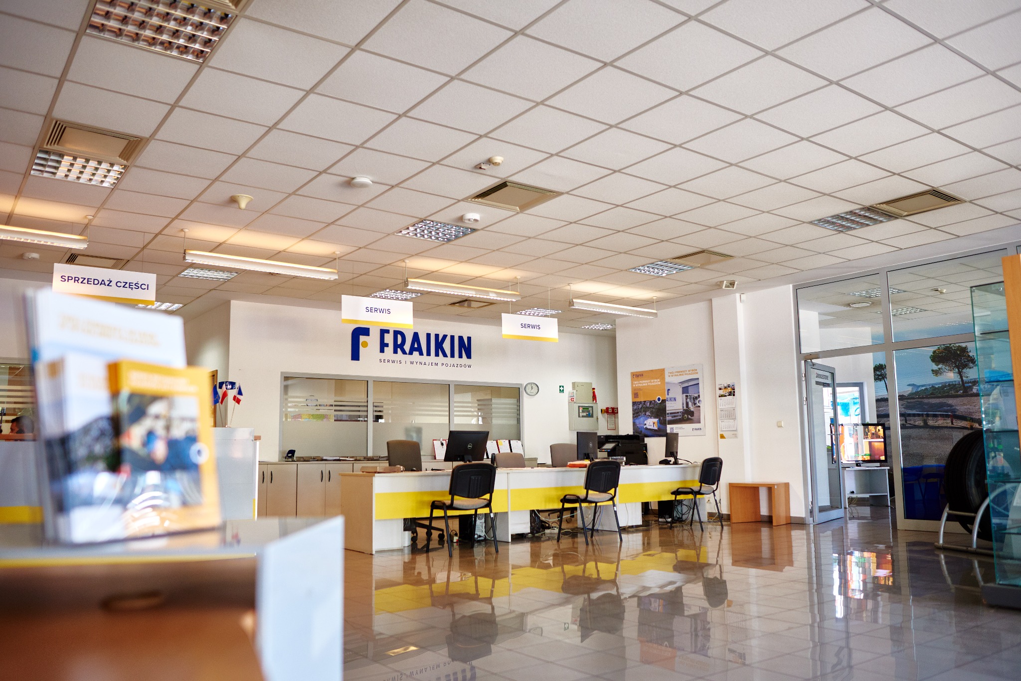 Jasne wnętrze punktu obsługi klienta serwisu samochodowego z widocznym biurkiem recepcyjnym, komputerami, krzesłami i logo firmy na ścianie. Na pierwszym planie ulotki reklamowe. Podłoga z połyskiem.