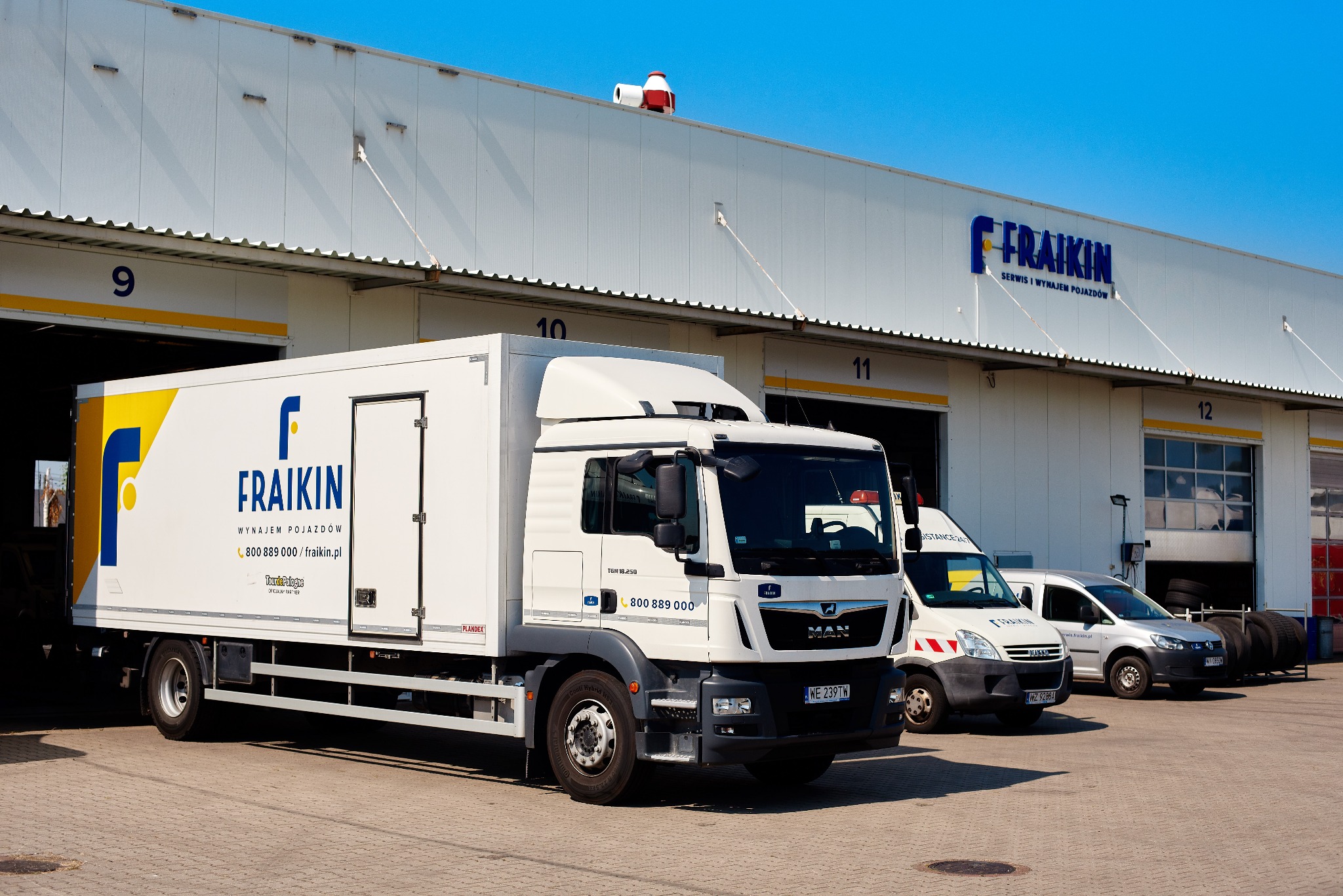 Ciężarówka z zabudową kontenerową marki MAN z logo Fraikin zaparkowana przed budynkiem serwisu i wynajmu pojazdów Fraikin, obok zaparkowane busy i samochody osobowe, widoczne numery bram wjazdowych.