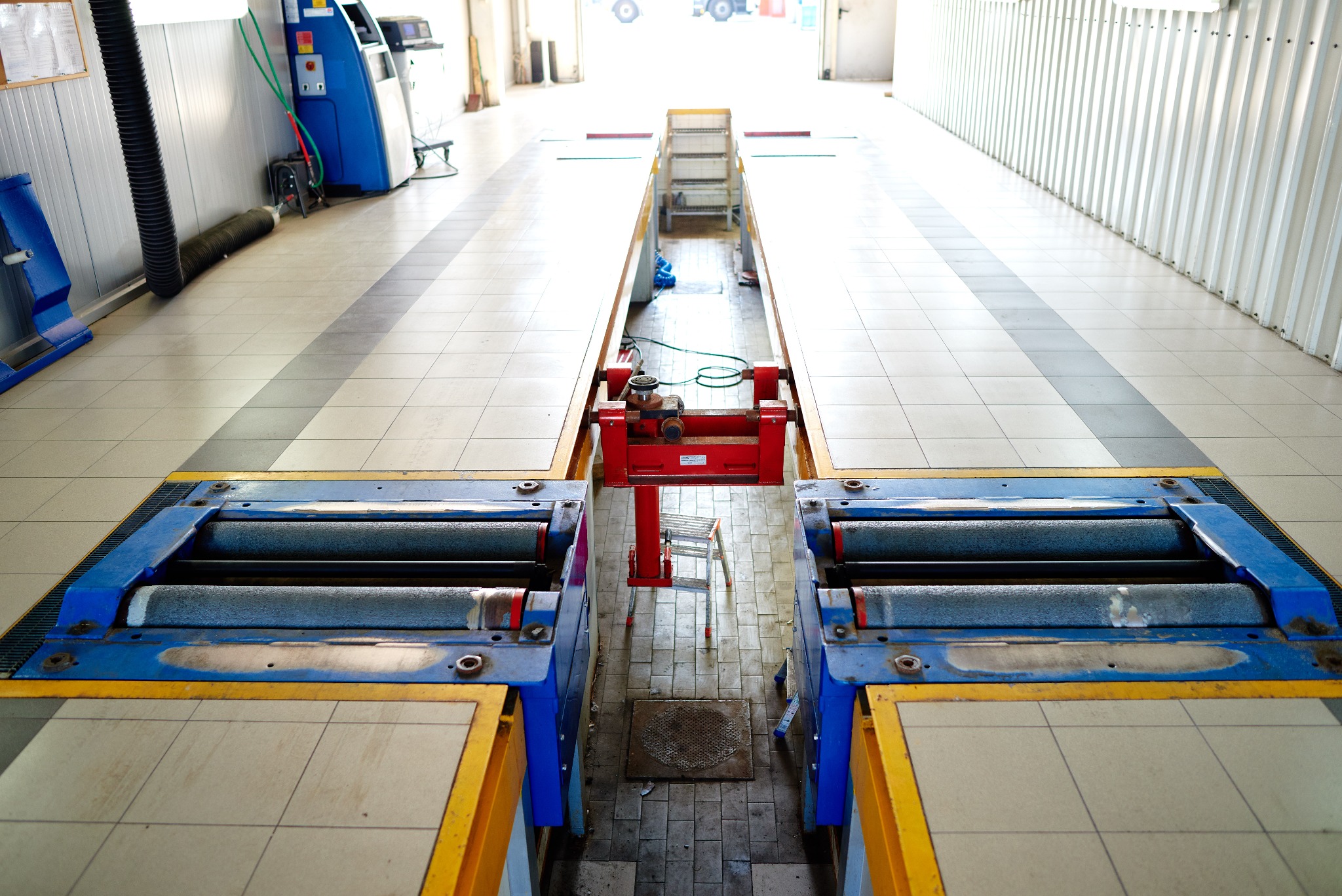 Stanowisko do testowania hamulców w warsztacie samochodowym z widocznym urządzeniem rolkowym, podnośnikiem i schodami. Posadzka wyłożona płytkami ceramicznymi.