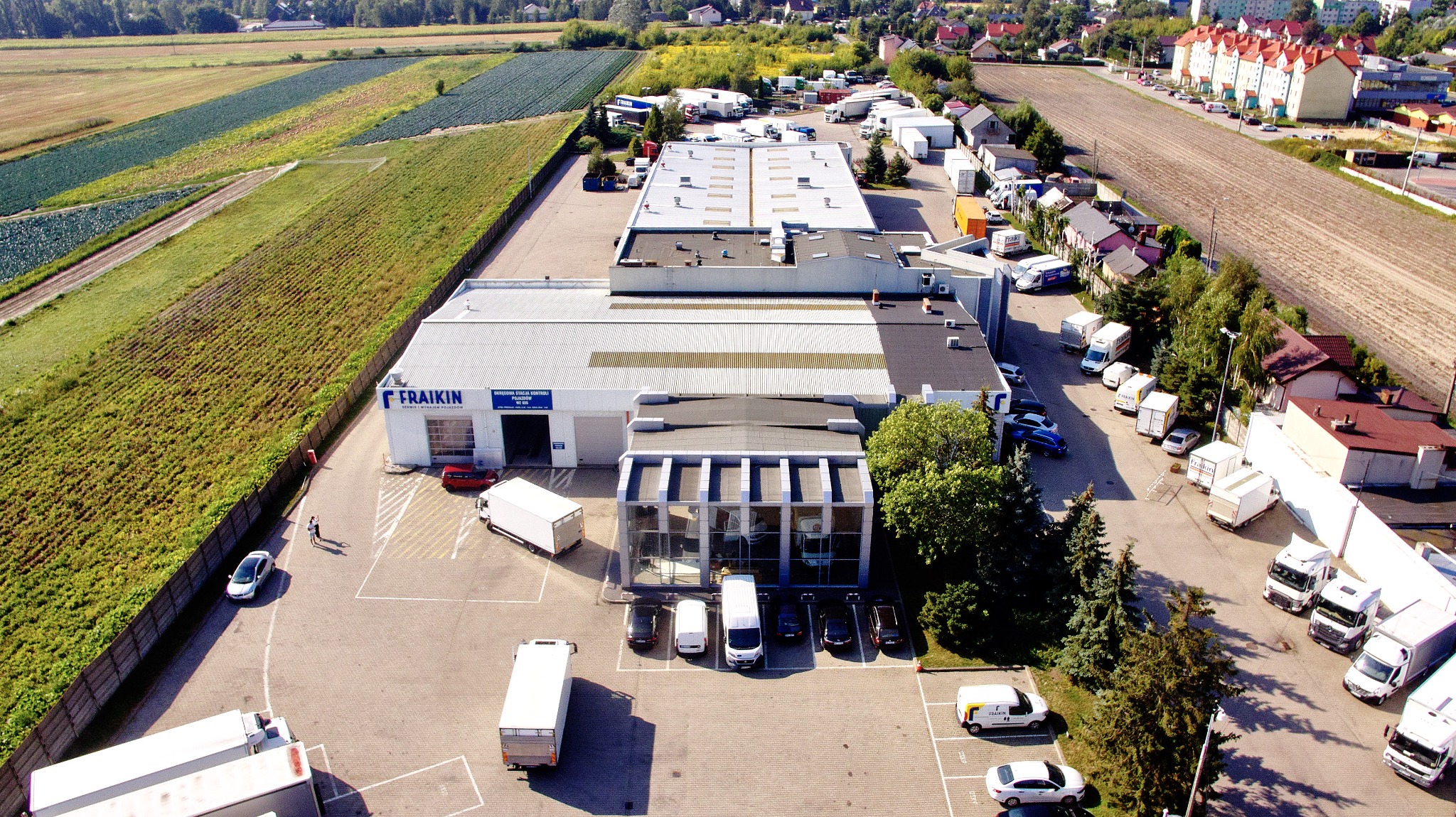 Widok z lotu ptaka na budynek firmy z zaparkowanymi busami i ciężarówkami, w tle pola uprawne i zabudowa mieszkalna.