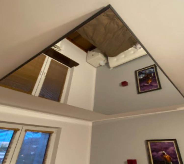 Trójkątne lustro na suficie odbija fragment pokoju z oknami w białych ramach, szarymi ścianami i obrazami w czarnych ramkach.