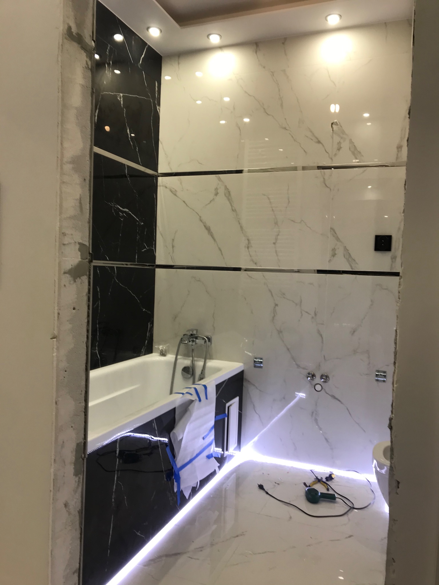 Remontowana łazienka z czarno-białymi płytkami imitującymi marmur, widoczne narzędzia na podłodze i fragment wanny z zabezpieczeniem folią.