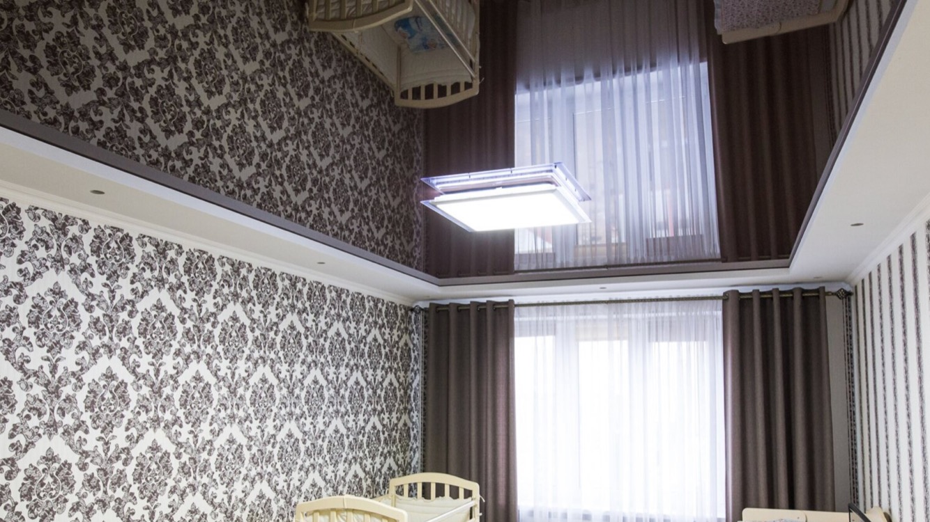 Pokój dziecięcy z wzorzystą tapetą, błyszczącym, ciemnobrązowym sufitem podwieszanym z wbudowanym oświetleniem LED, jasnymi firanami i ciemnymi zasłonami oraz białym łóżeczkiem niemowlęcym.