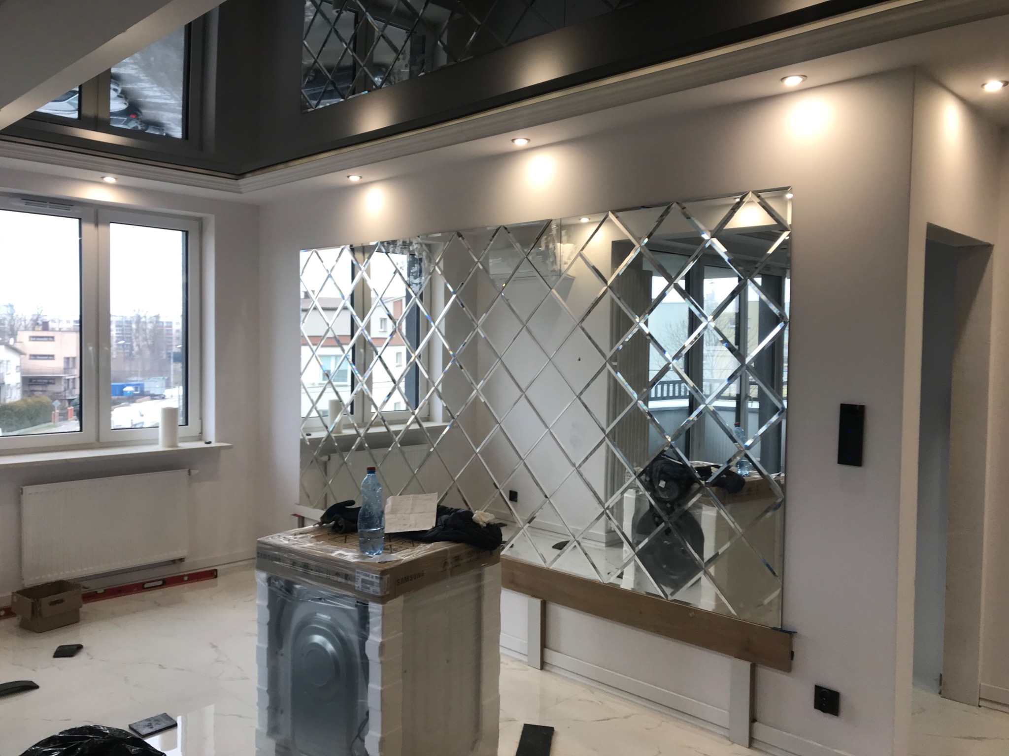 Ściana z dekoracyjnych, romboidalnych luster w jasnym pokoju, widoczny nowy sprzęt AGD w opakowaniu i odbicie okna w lustrach.