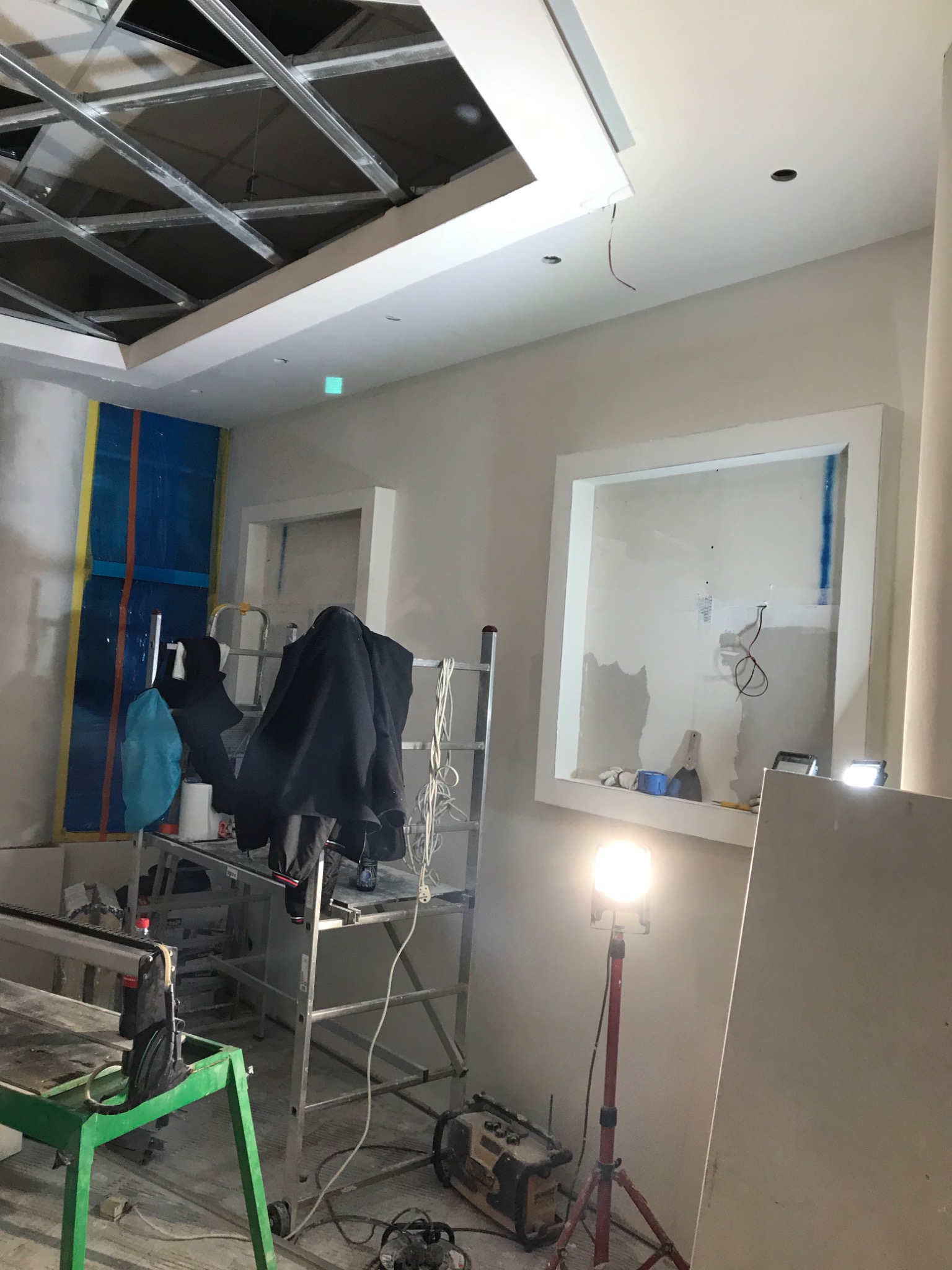 Wnętrze w trakcie prac wykończeniowych: widoczna konstrukcja sufitu podwieszanego, wnęka w ścianie, drabina z ubraniami, narzędzia i lampa budowlana.