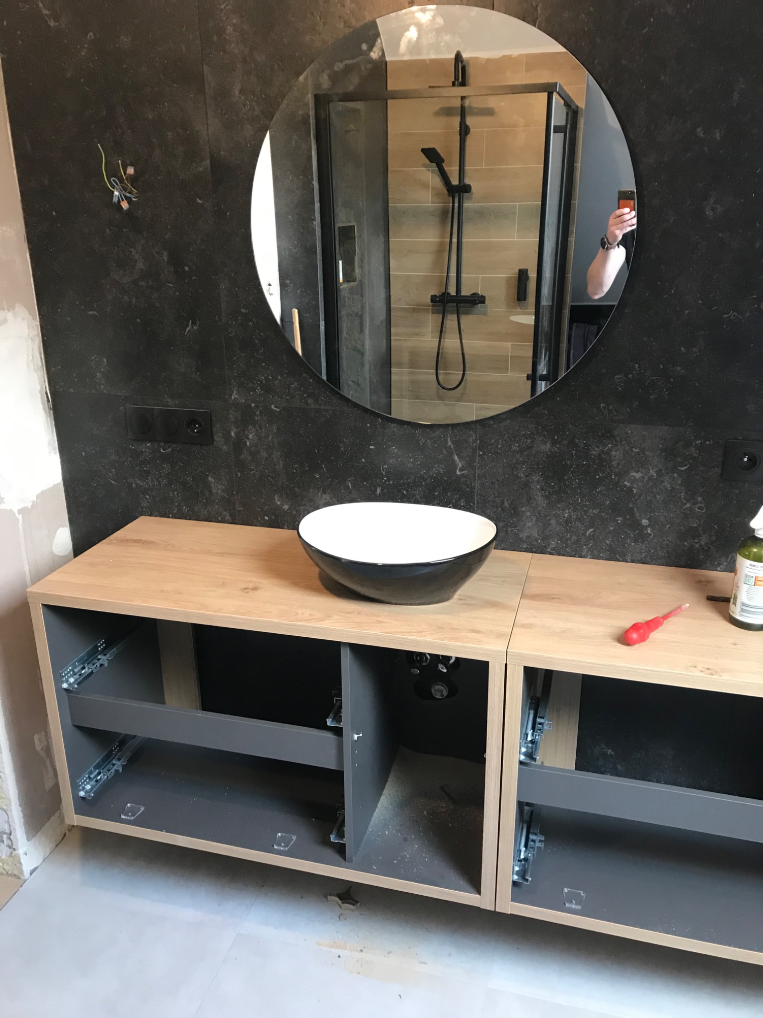 Łazienka w trakcie wykańczania: montaż szafki pod umywalkę z owalną, czarną misą umywalkową, na ścianie ciemne płytki imitujące kamień, okrągłe lustro odbijające kabinę prysznicową.