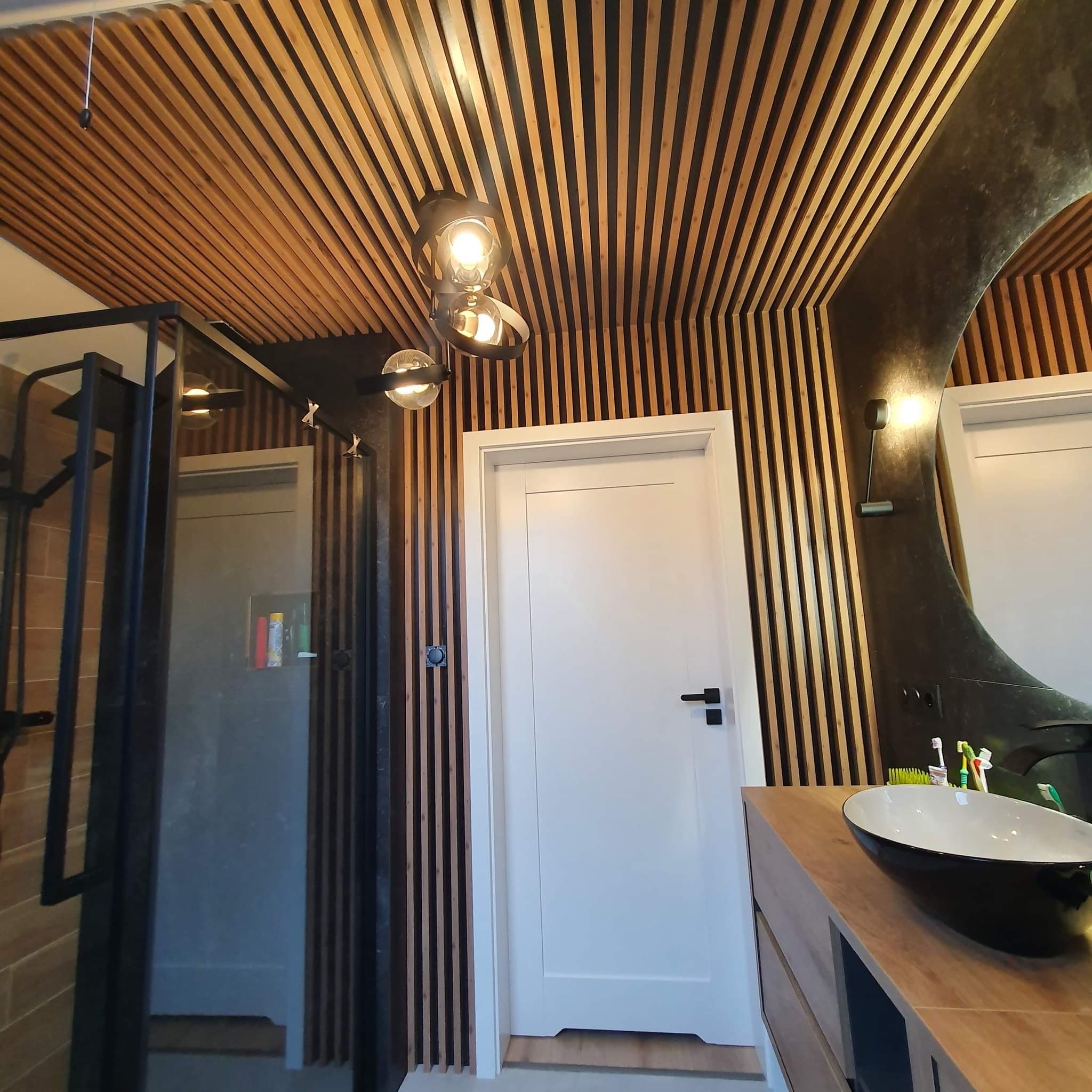 Nowoczesna łazienka z drewnianymi lamelami na ścianach i suficie, okrągłe lustro z podświetleniem, czarna umywalka nablatowa i kabina prysznicowa z czarnymi profilami.