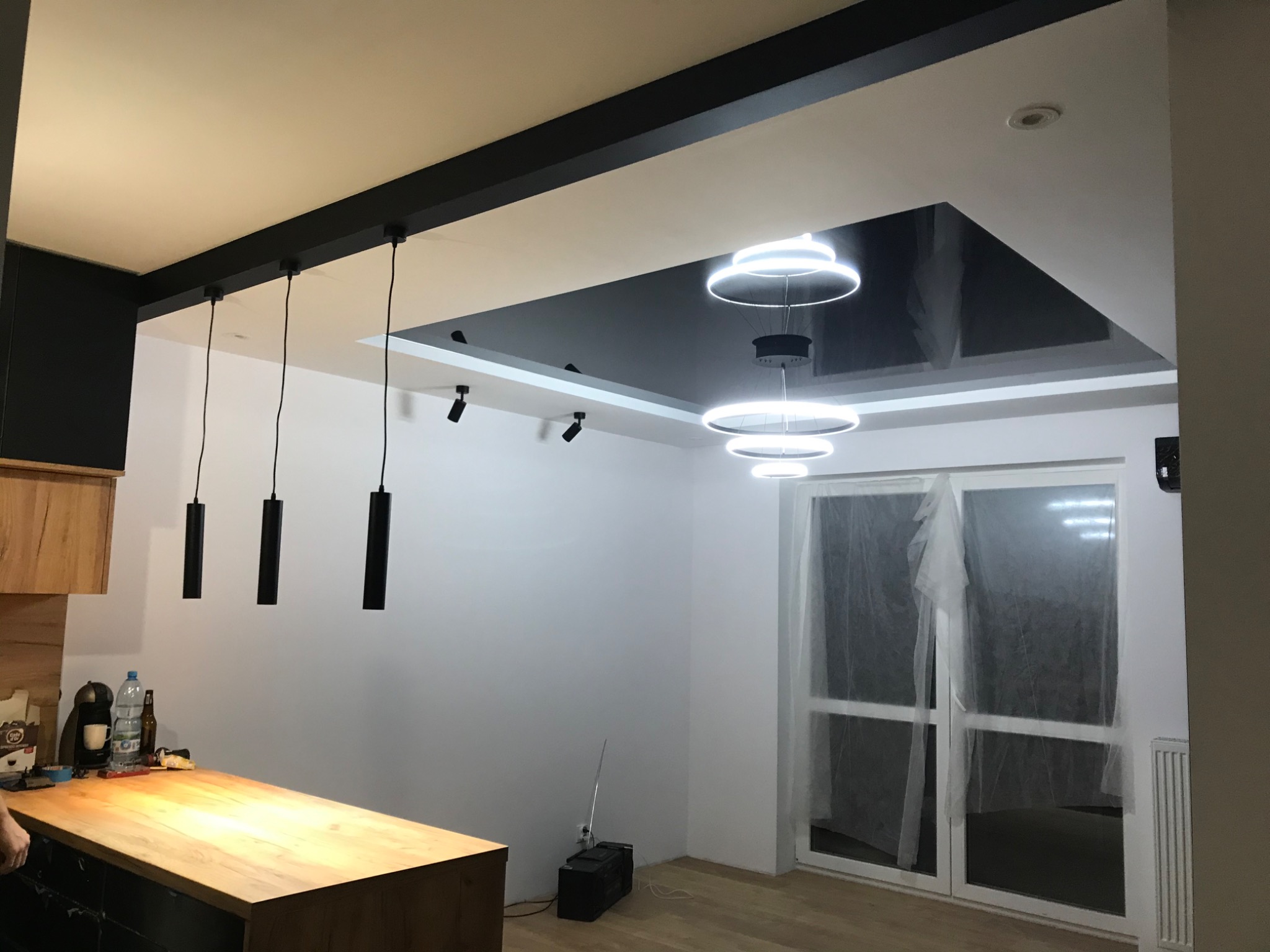 Nowoczesne oświetlenie w salonie z czarnym, błyszczącym sufitem podwieszanym, okrągłe lampy LED i wiszące cylindryczne lampy nad drewnianym blatem kuchennym.