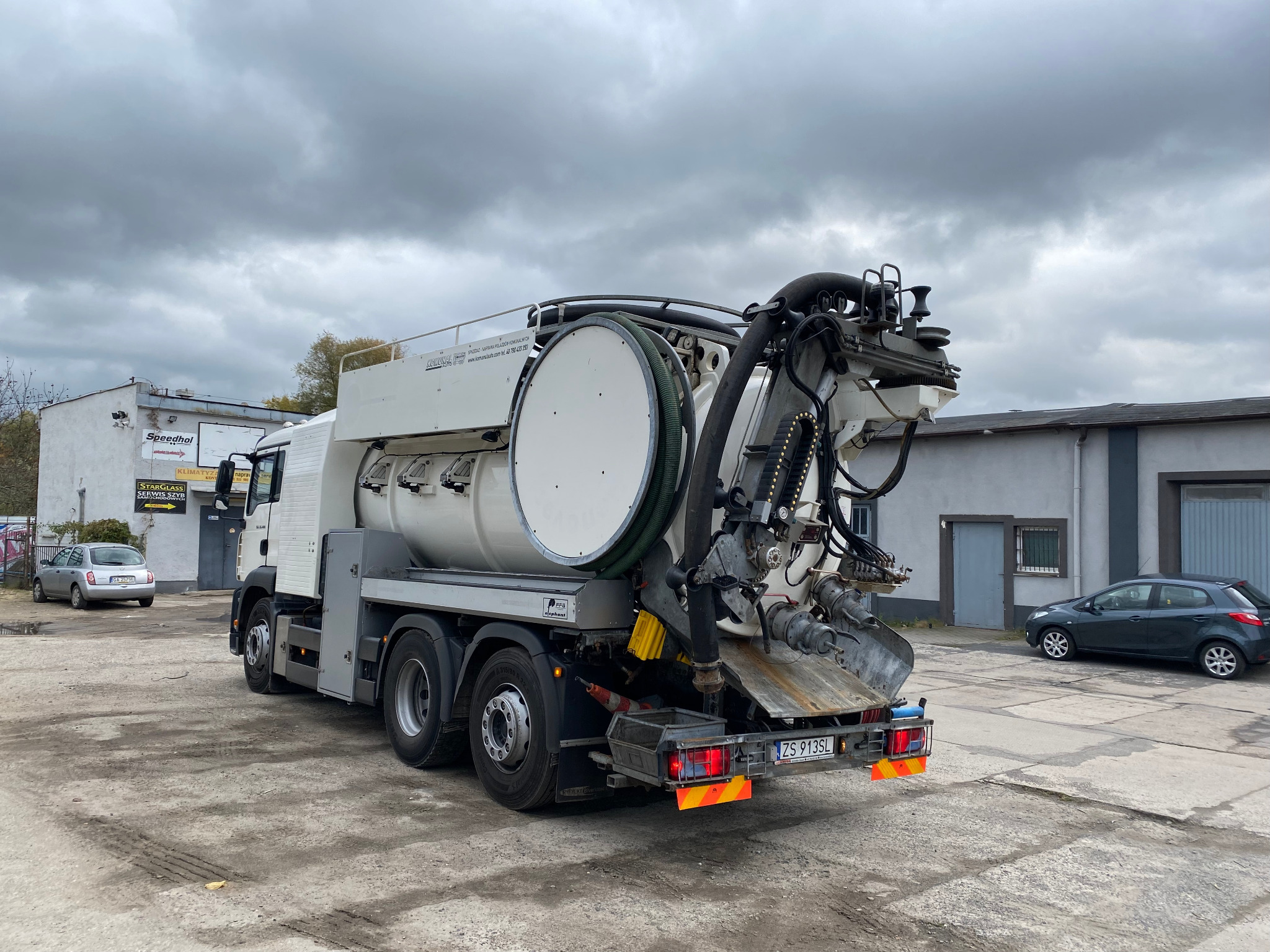 Pojazd asenizacyjny z dużym zbiornikiem i systemem rur ssących, zaparkowany na placu w Stargardzie; widoczny budynek z szyldem Speedhol w tle.