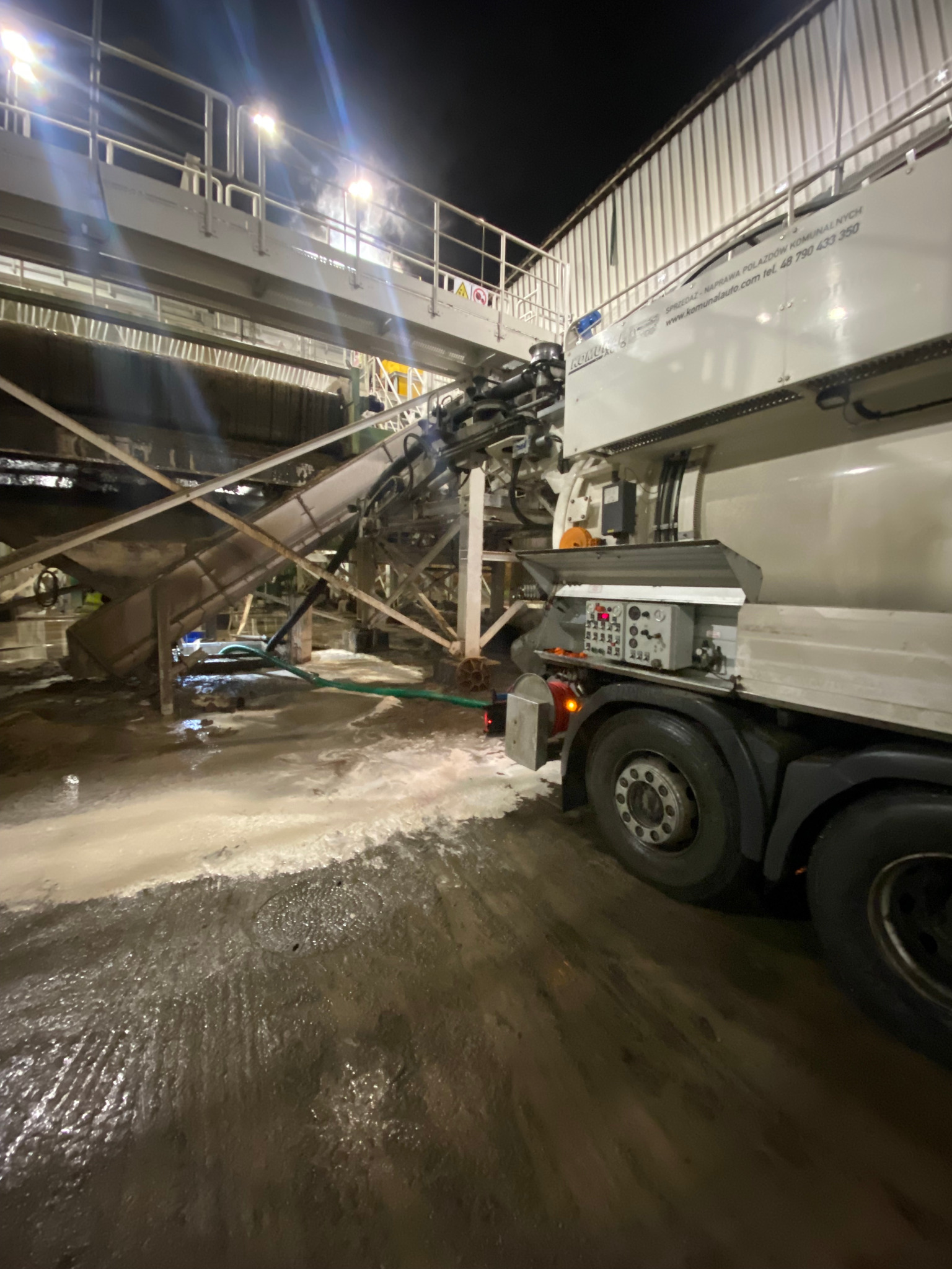 Specjalistyczna ciężarówka z podnośnikiem i systemem czyszczenia pod ciśnieniem, z zielonym wężem, w trakcie pracy na zewnątrz budynku przemysłowego, widać mokrą powierzchnię po czyszczeniu.