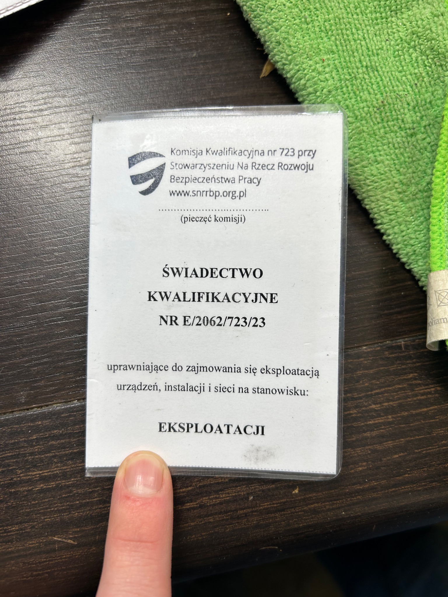 Świadectwo Kwalifikacyjne nr E/2062/723/23 uprawniające do zajmowania się eksploatacją urządzeń, instalacji i sieci, leżące na ciemnym drewnianym stole obok zielonej ściereczki, wskazane palcem