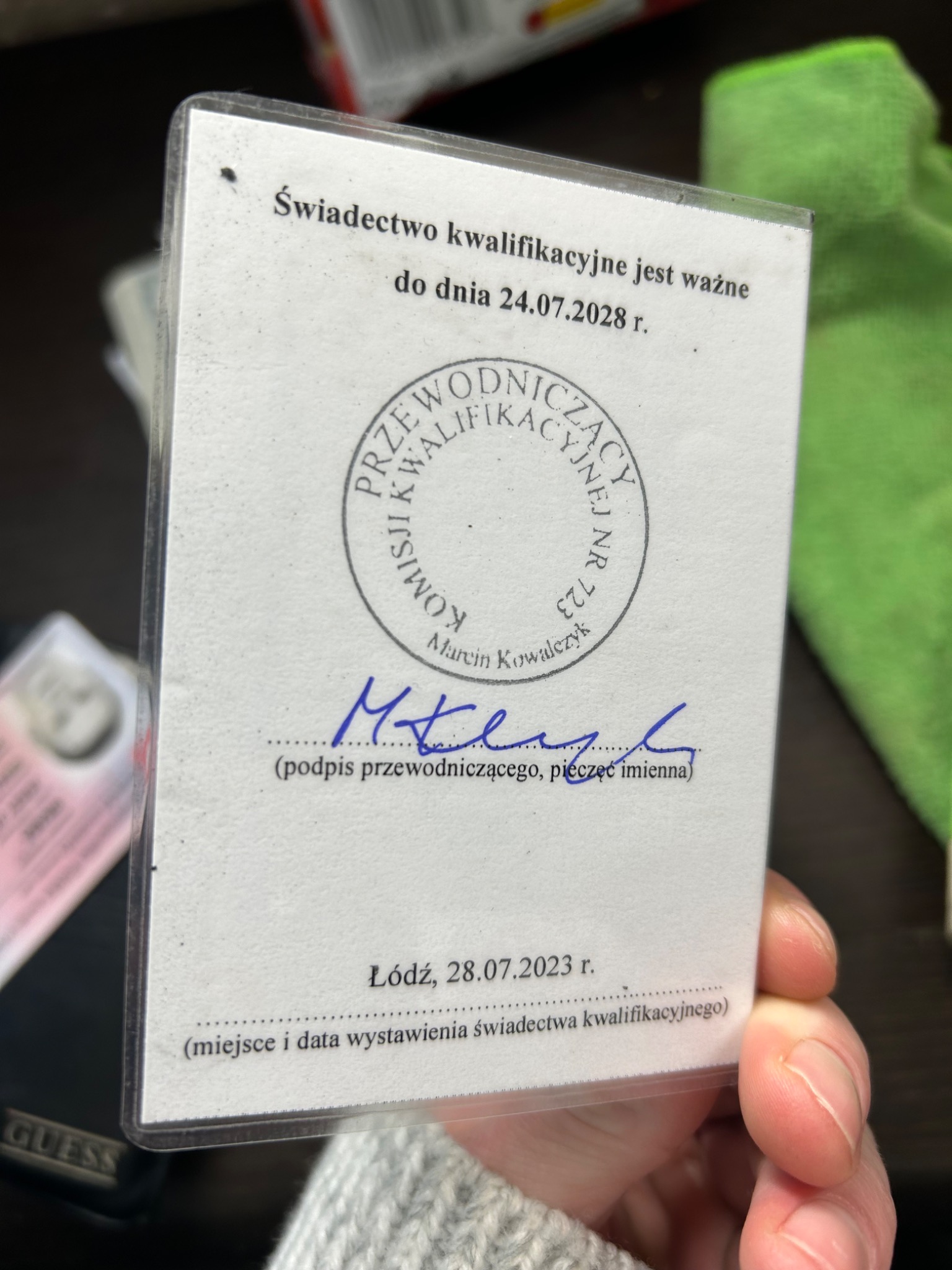 Świadectwo kwalifikacyjne z pieczęcią i podpisem, ważne do 24.07.2028, trzymane w ręce na rozmytym tle.