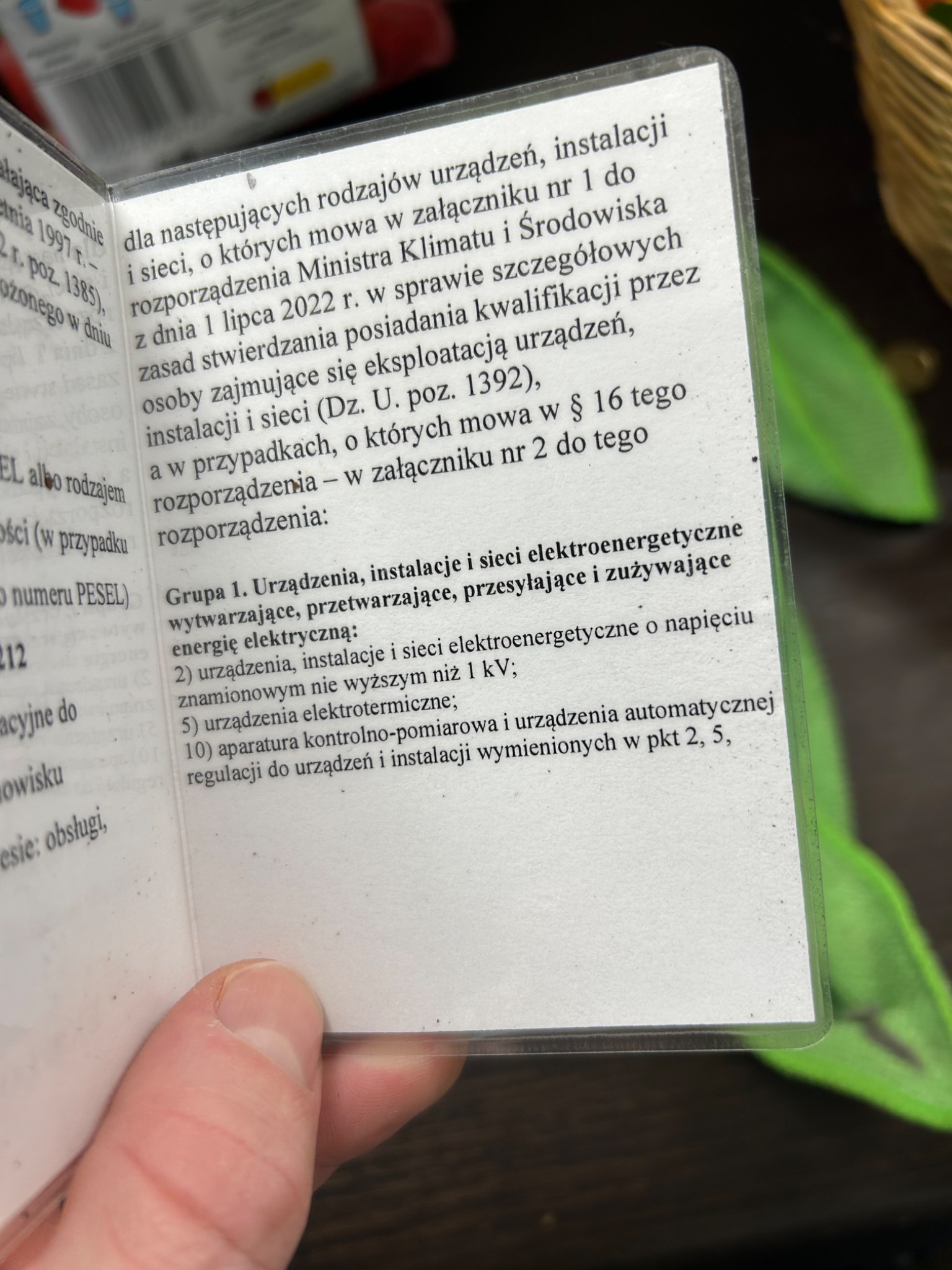 Zbliżenie na otwartą legitymację z tekstem dotyczącym uprawnień do eksploatacji urządzeń, instalacji i sieci elektroenergetycznych, trzymaną w dłoni, z widocznymi fragmentami przepisów i regulacji.
