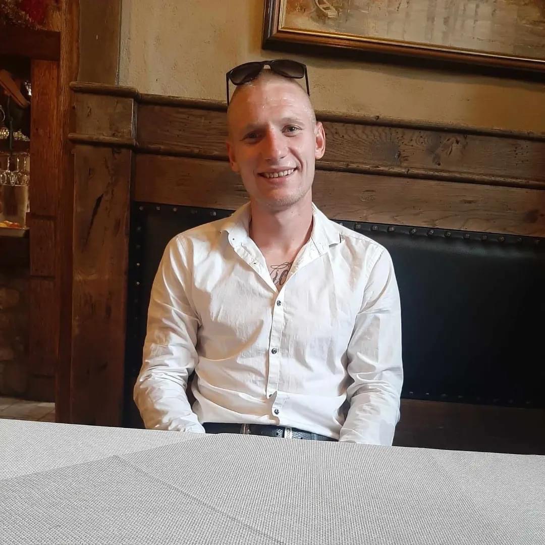 Uśmiechnięty mężczyzna w białej koszuli siedzi przy stole w rustykalnym wnętrzu z drewnianymi panelami i ciemną skórzaną ławką w tle, okulary przeciwsłoneczne na głowie.