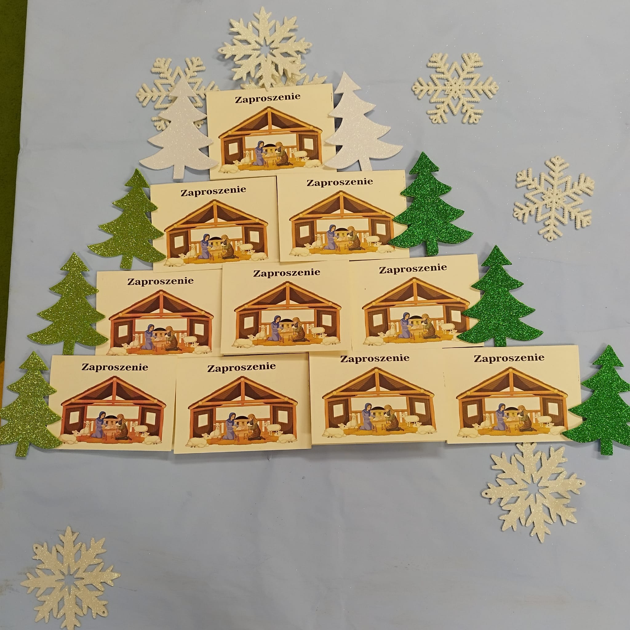 Stos kartek z napisem 'Zaproszenie' i ilustracją szopki bożonarodzeniowej, otoczony dekoracjami świątecznymi: brokatowymi choinkami i śnieżynkami, ułożony na jasnoniebieskim tle.