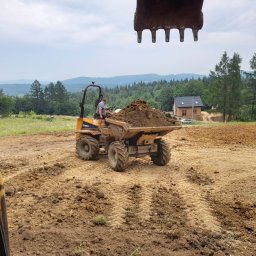 Firma Handlowo Usługowo Transportowa Krystian Bieniek - Żółta wywrotka z ładunkiem ziemi na placu budowy, widok z kabiny koparki, w tle dom jednorodzinny i zalesione wzgórza.